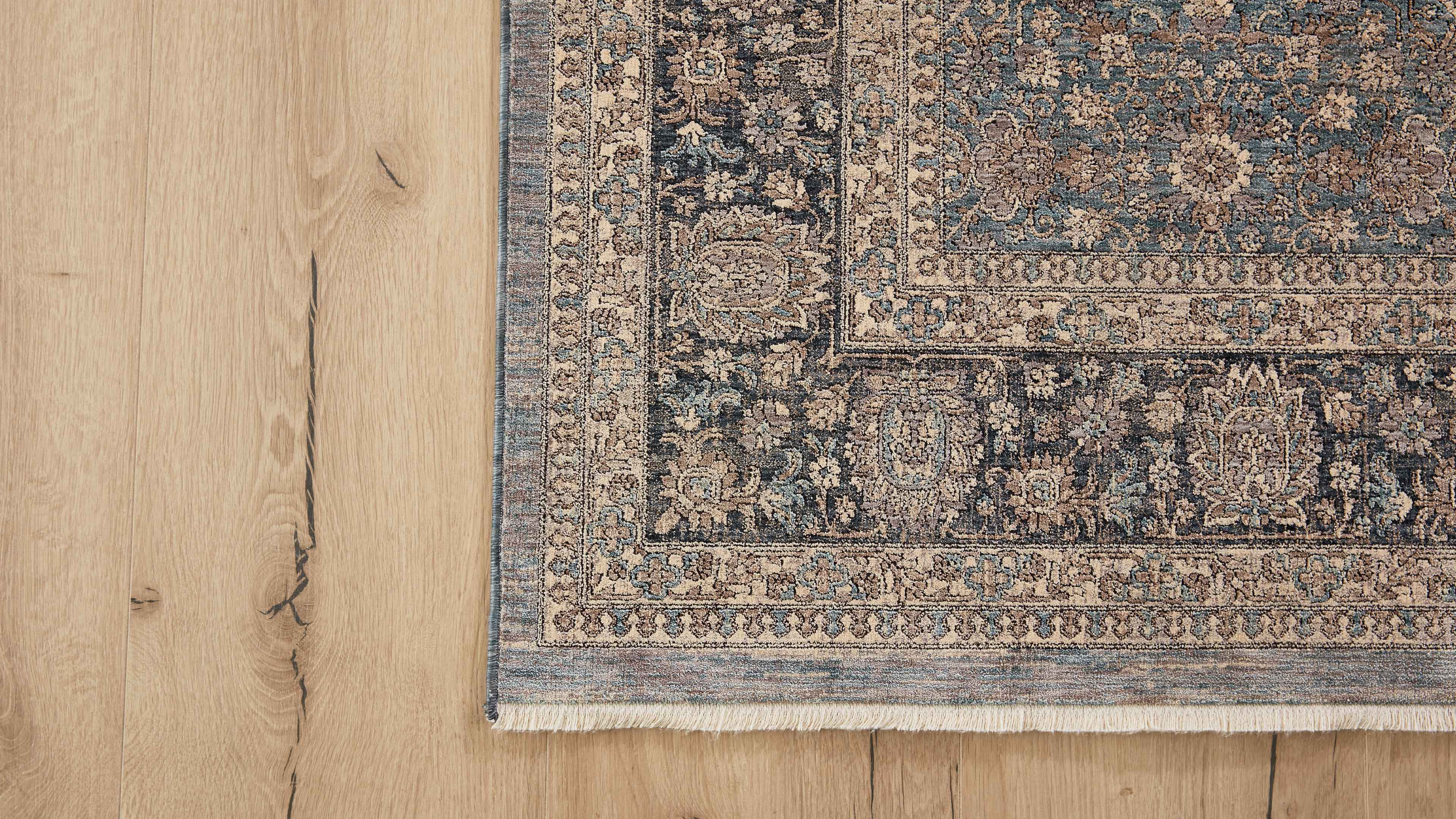 Eckansicht eines gemusterten Teppichs mit Fransen und floralen Ornamenten in Beige, Blau und Braun auf hellem Holzfußboden.