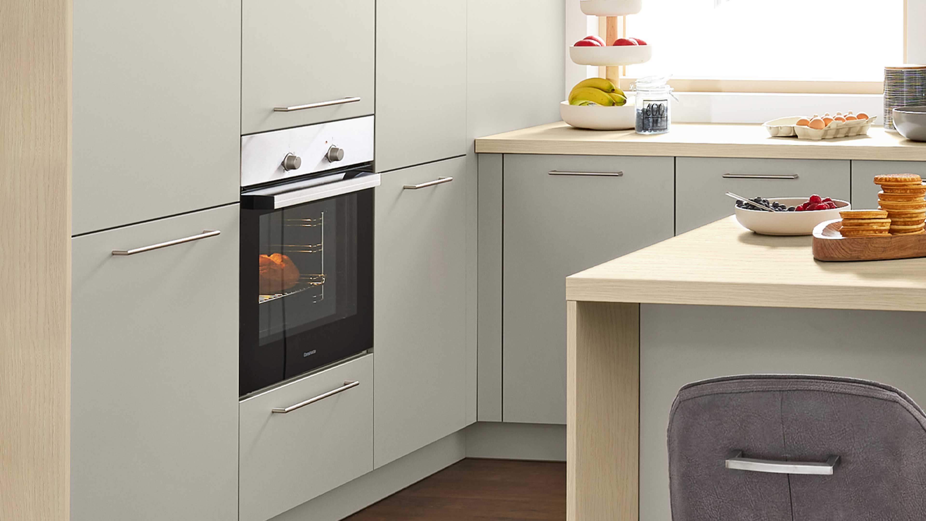 Eckküchenzeile mit hellgrauen Fronten und Edelstahlgriffen, eingebautem schwarzen Ofen mit Backgut, Holzarbeitsplatte und grauem Stuhl.
