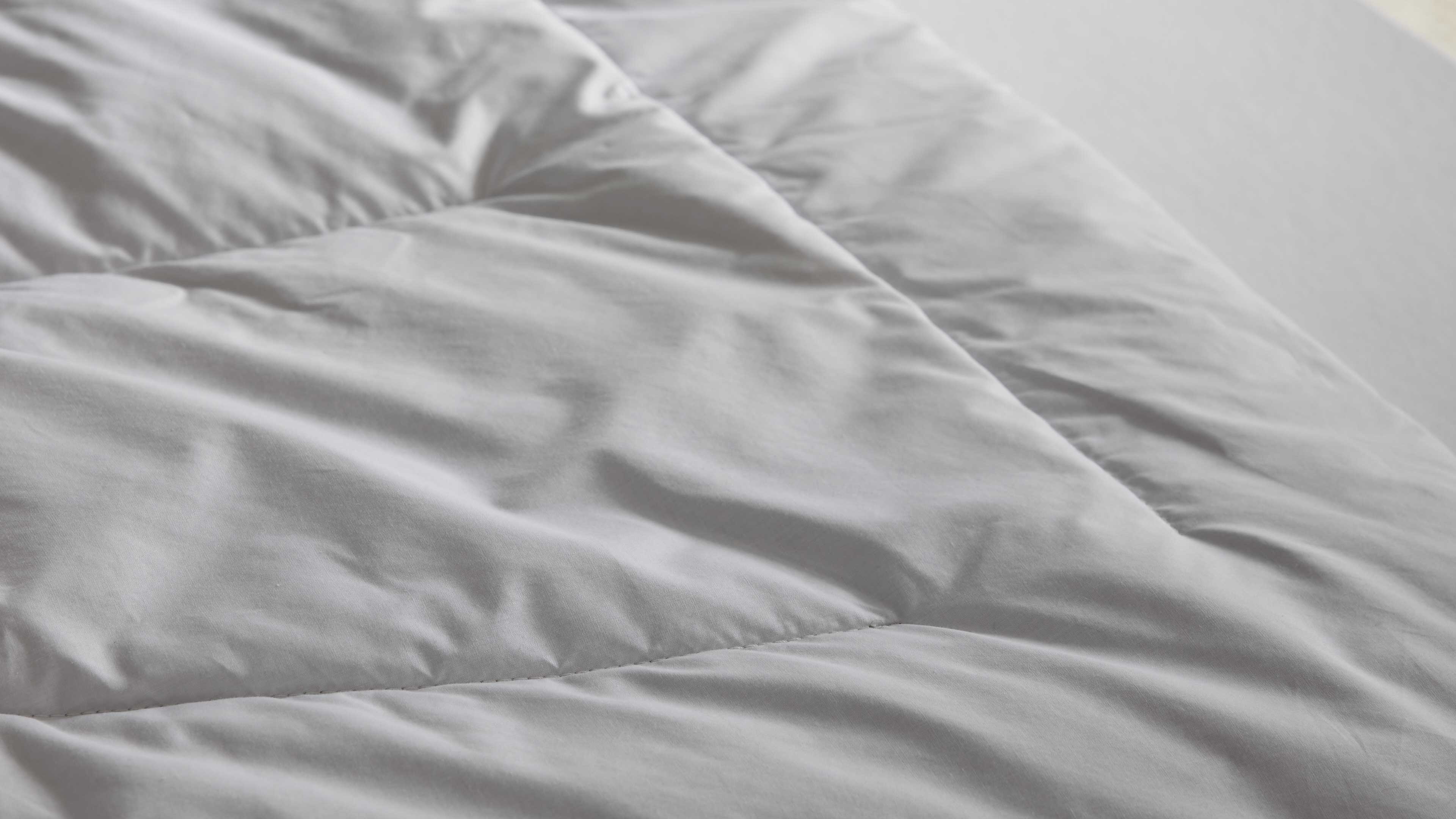 Weiße, gesteppte Bettdecke mit rechteckigen Nähten und leicht gewellter Oberfläche, auf hellem Matratzenbezug liegend.