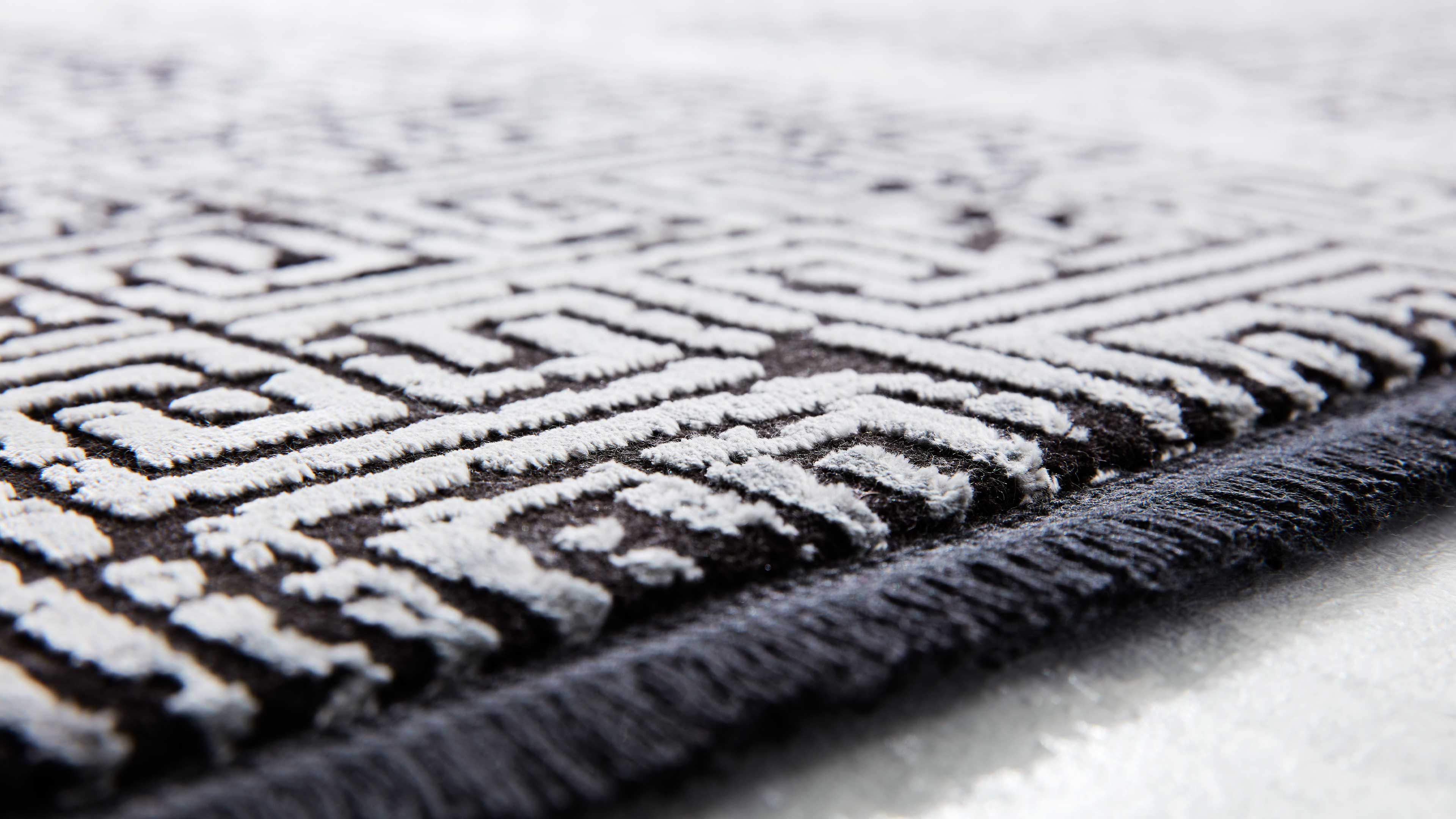 Nahaufnahme eines dunkelgrauen Teppichs mit weißem, erhabenem geometrischen Labyrinthmuster, dunkler Kettelung und sichtbarer Florstruktur.