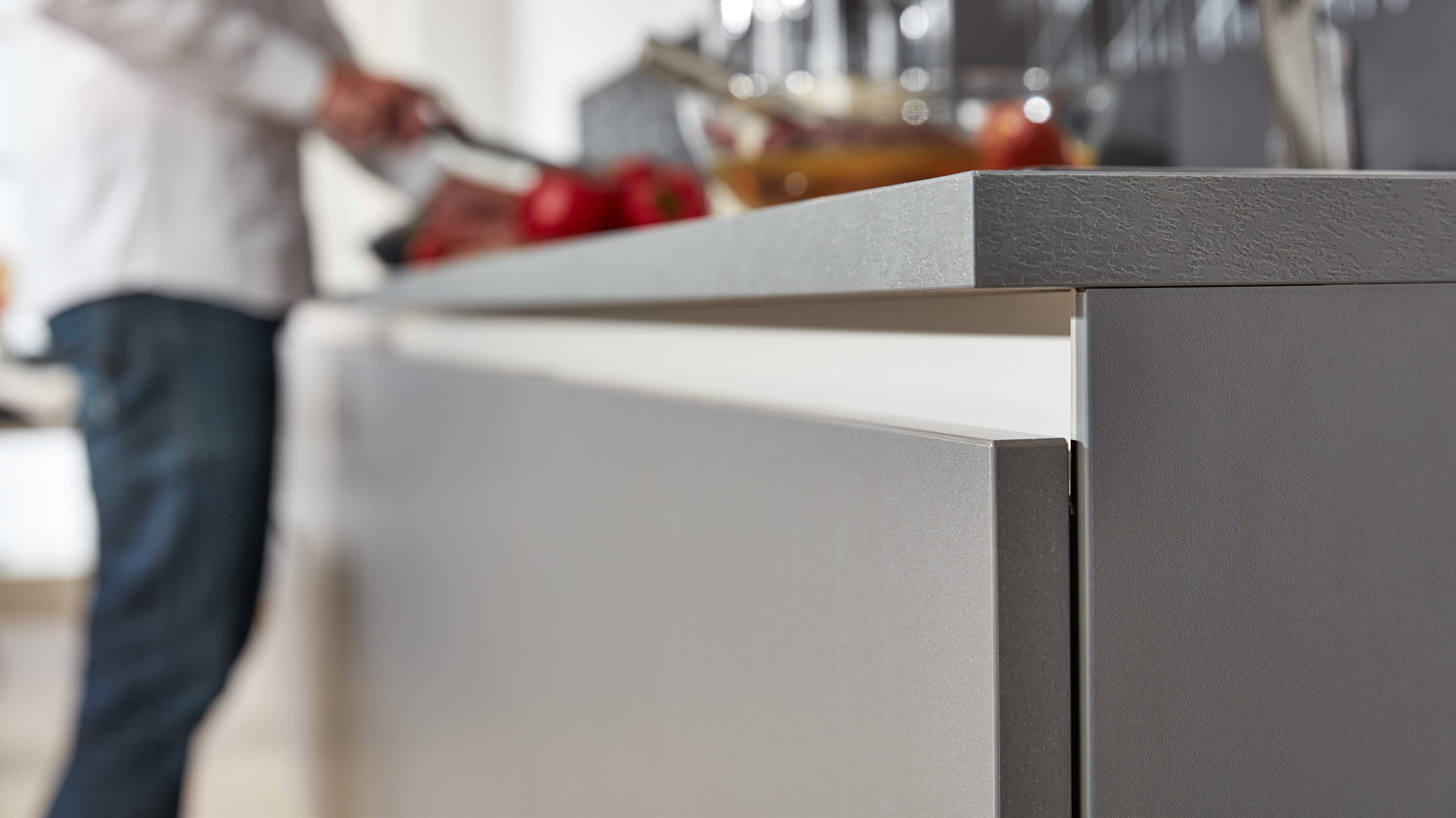 Nahaufnahme einer grifflosen Küchenfront in mattem Grau mit schmaler Schattenfuge und strukturiert wirkender heller Arbeitsplatte.