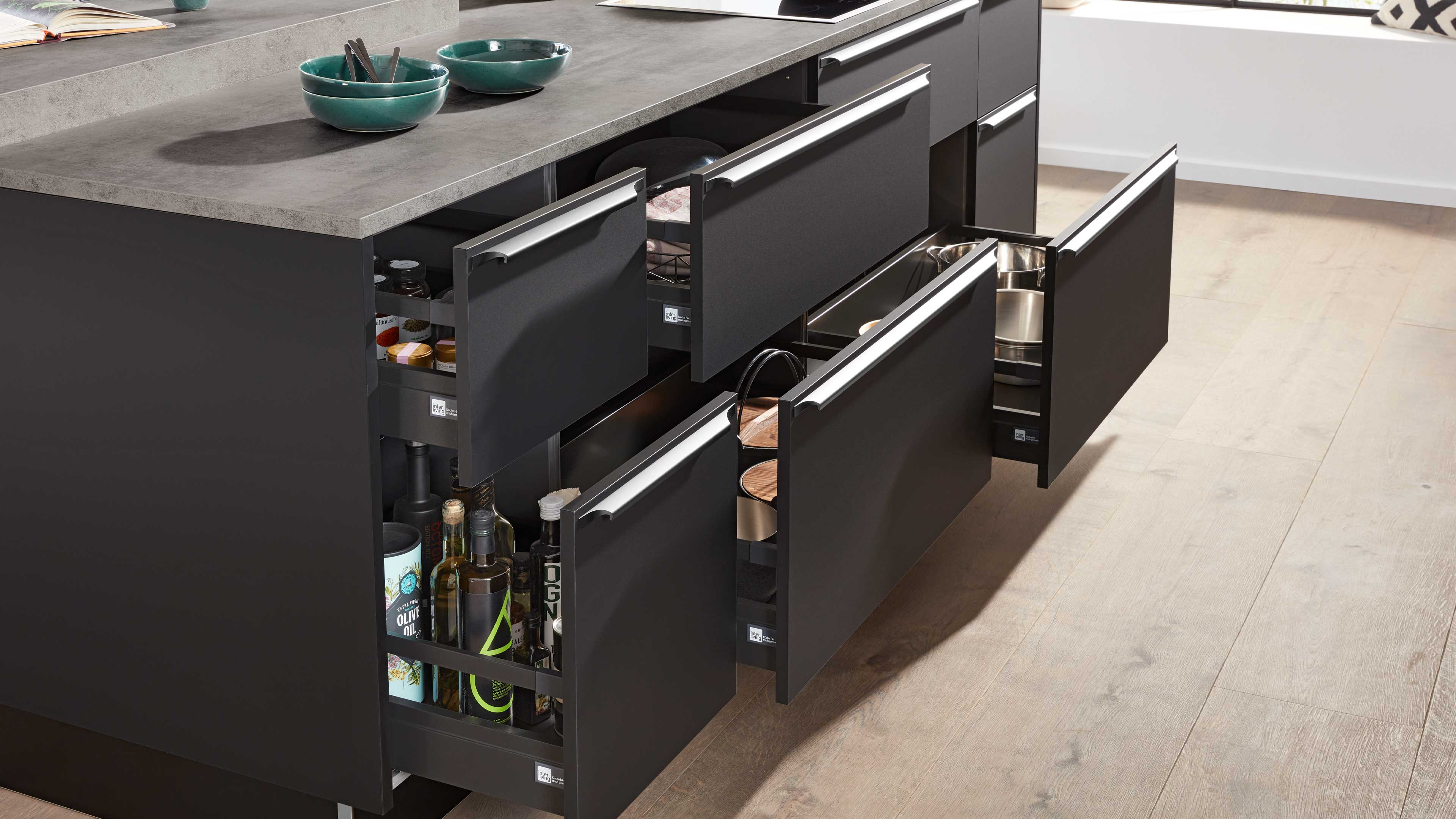 Offene dunkelgraue Küchenschubladen einer Kücheninsel mit herausgezogenen Auszügen, gefüllt mit Flaschen, Schüsseln und Töpfen.