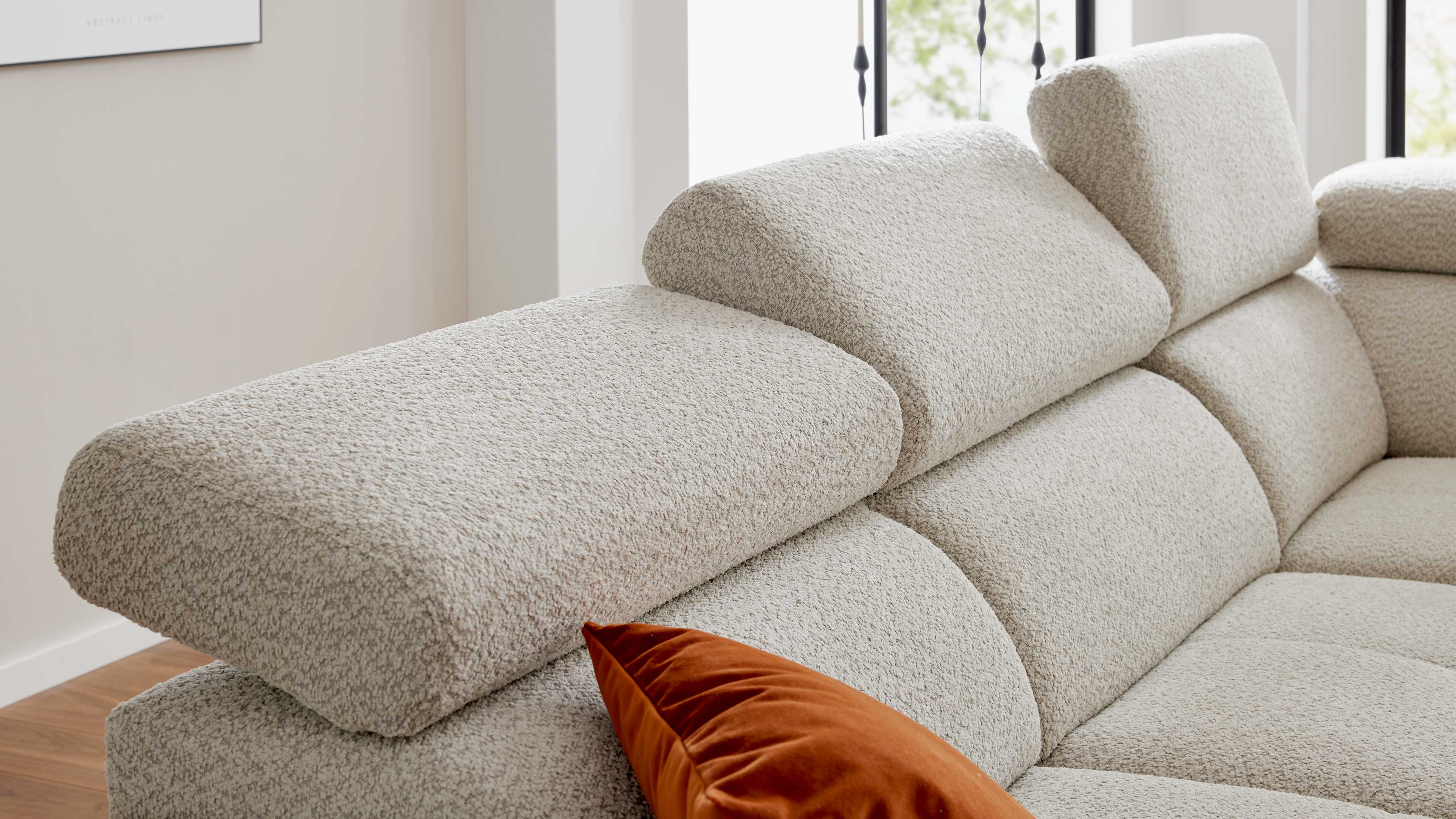 Beiges, strukturiertes Sofa mit verstellbaren Kopfstützen und orangefarbenem Samtkissen, seitlicher Fensterblick.