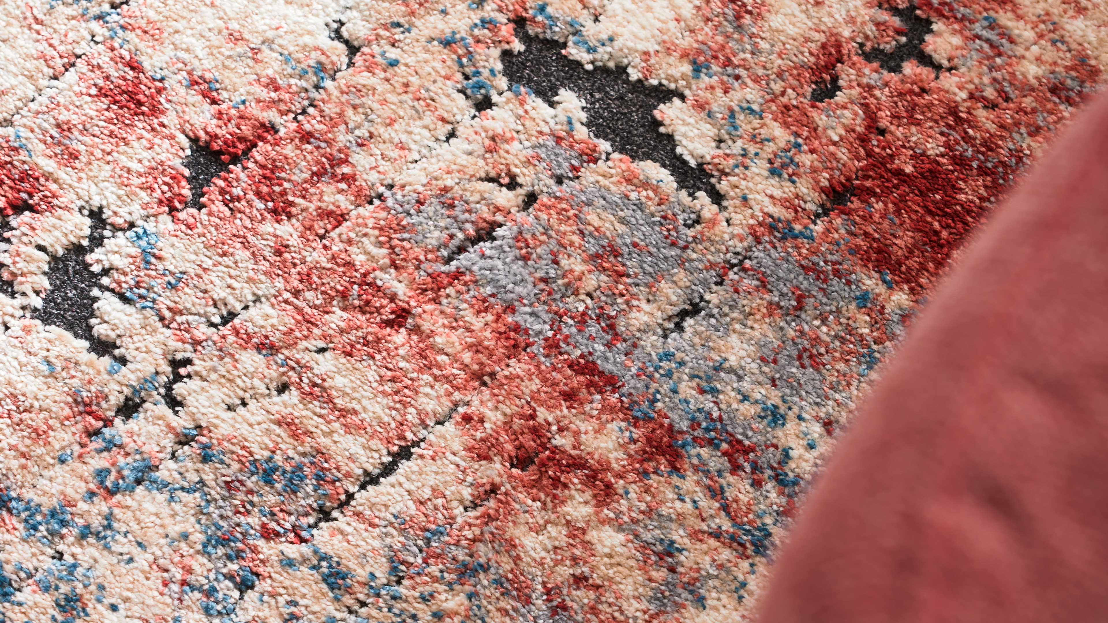 Nahaufnahme eines flauschigen Teppichs mit cremefarbenen, roten, blauen und grauen Flecken sowie schwarzen Linien, rechts rosafarbener Stoff.