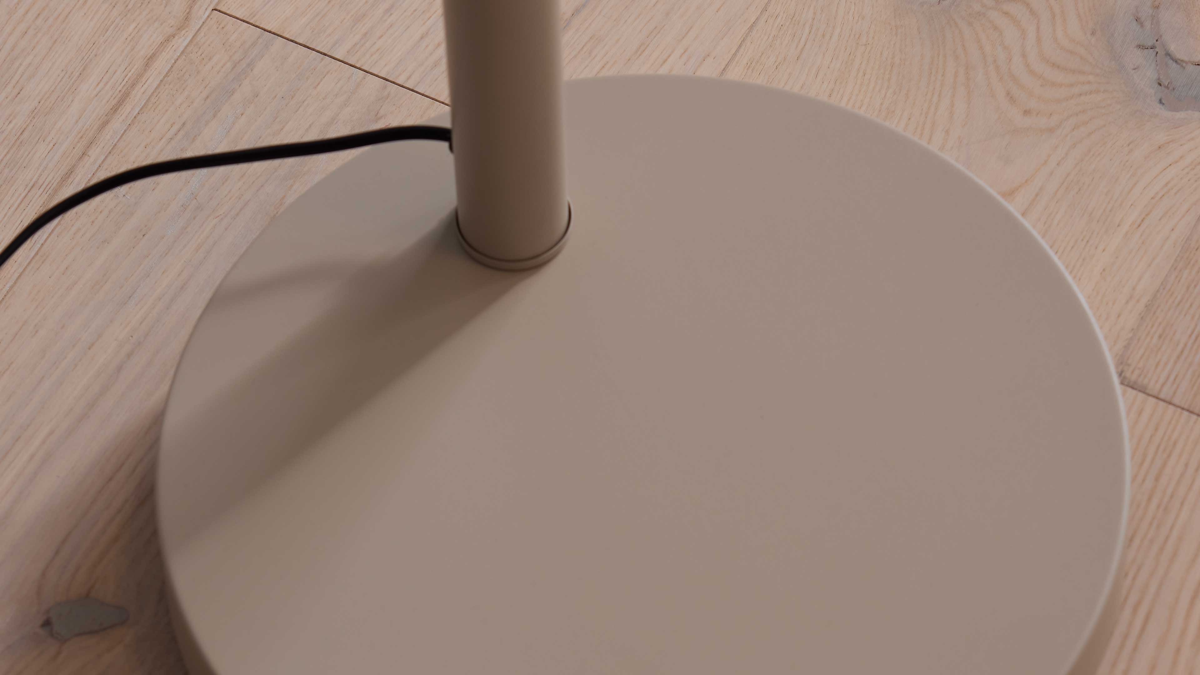 Runder mattbeiger Lampenfuß mit zylindrischem Stiel und schwarzem Kabel auf hell gemasertem Holzfußboden.