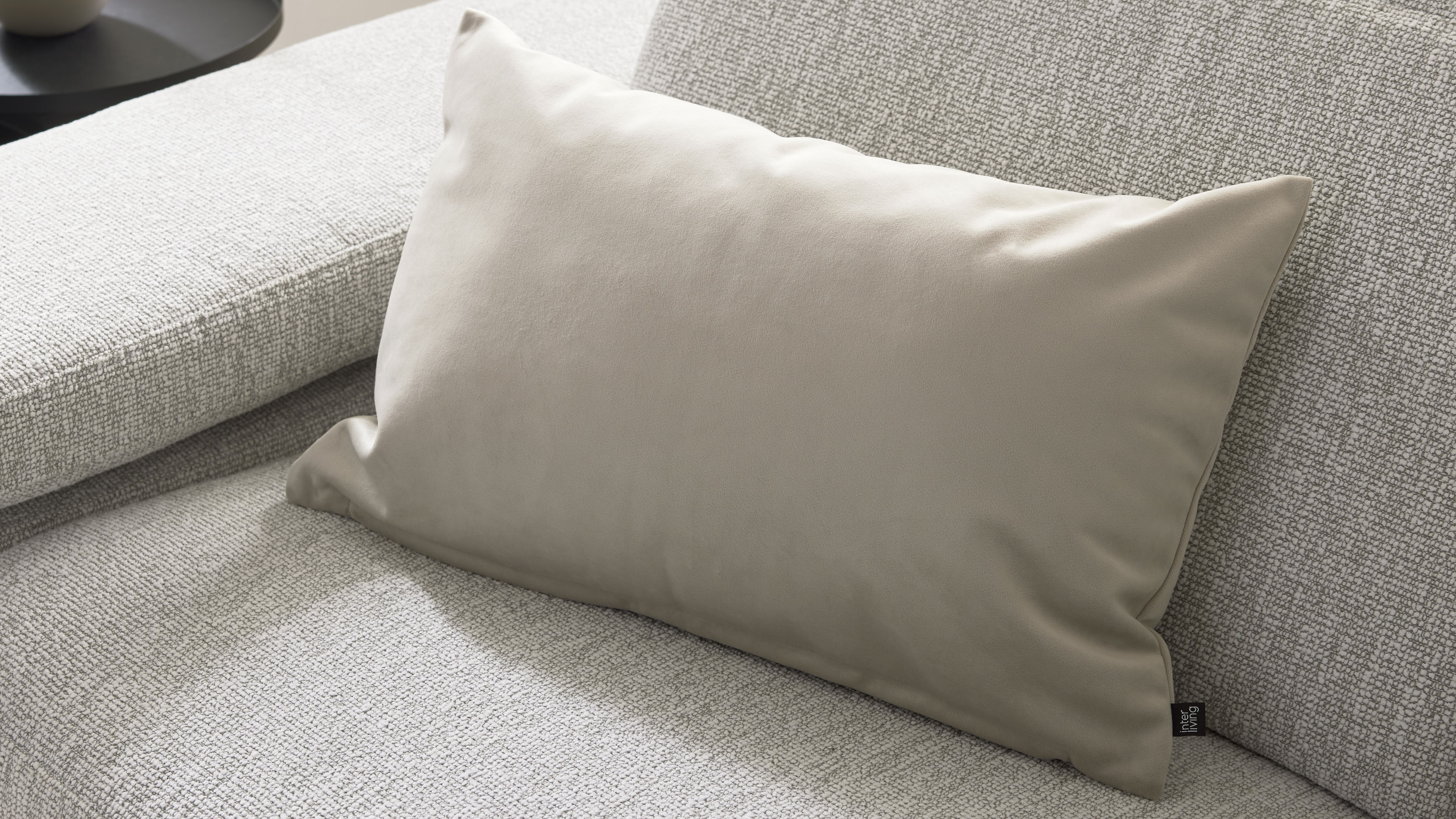 Rechteckiges beige Sofa-Kissen liegt auf hellgrauer, grob gewebter Sofafläche, rechts mit schwarzem Textiletikett.