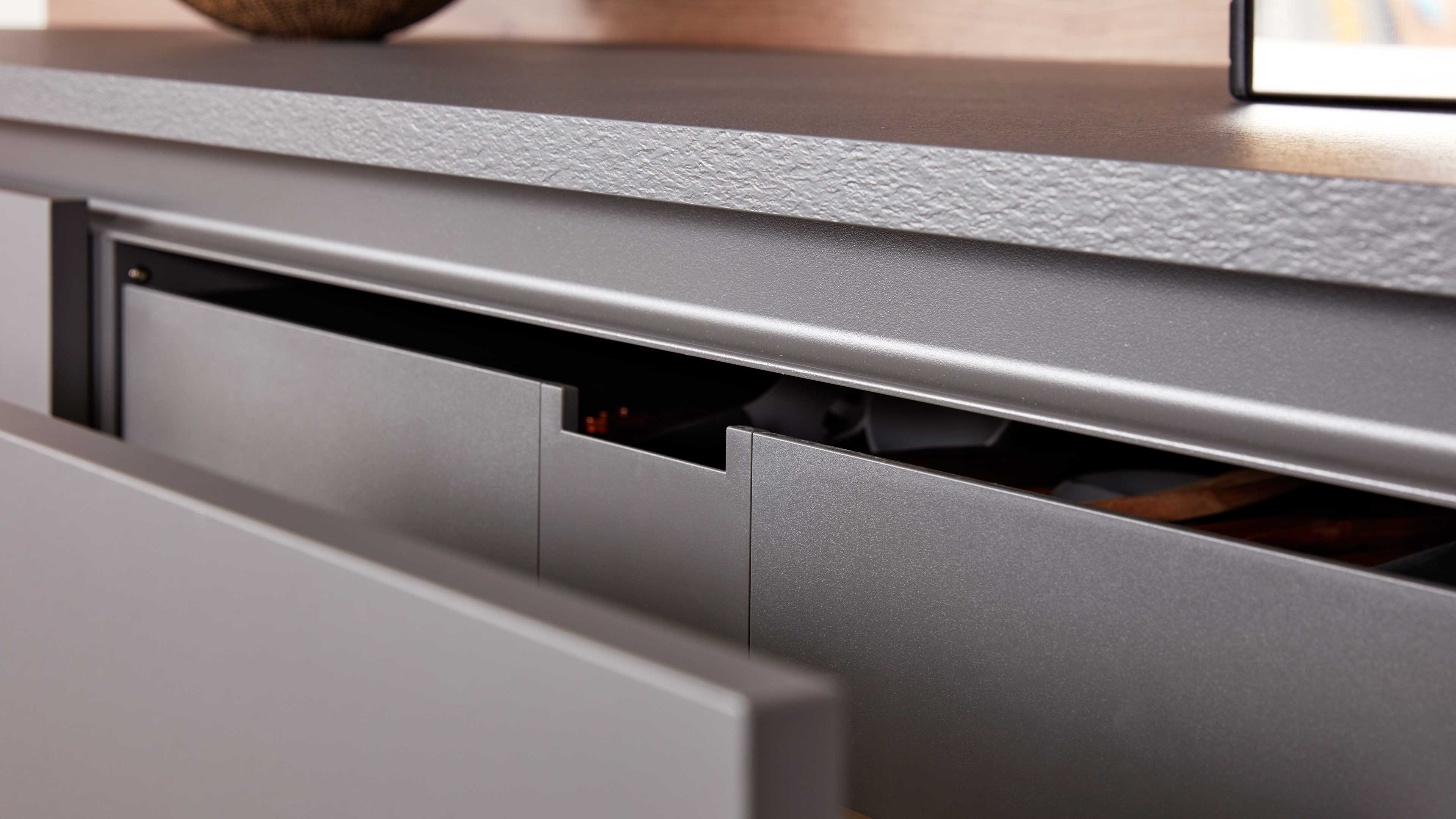 Graue, matt beschichtete Schubladenfronten mit eingekerbten Griffen unter einer rauen hellgrauen Arbeitsplatte, teilweise geöffnet.