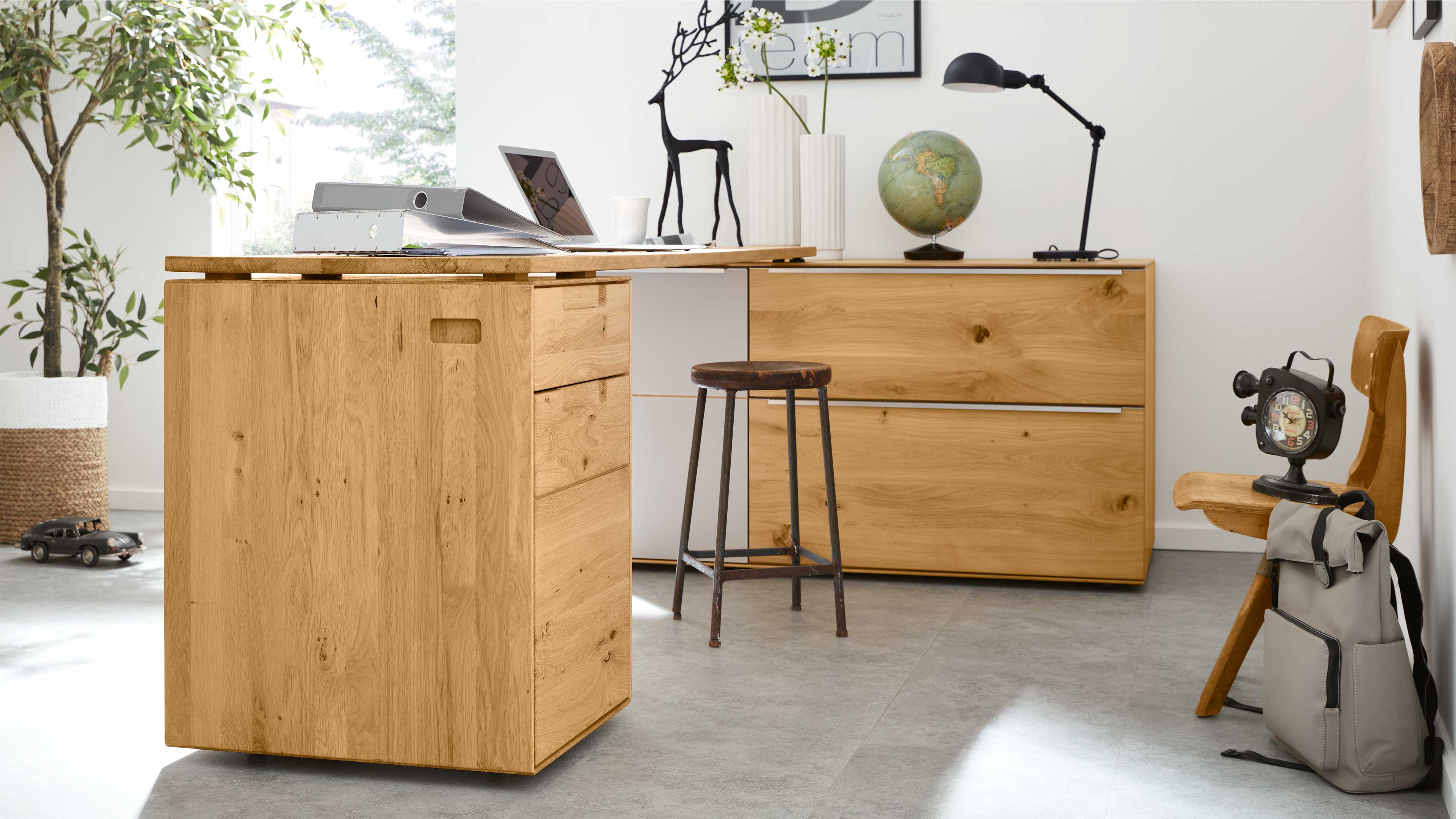 L-förmiger Schreibtisch aus hellem Holz mit Schubladenpodest links und Sideboard rechts, davor Hocker, Lampe und Globus.