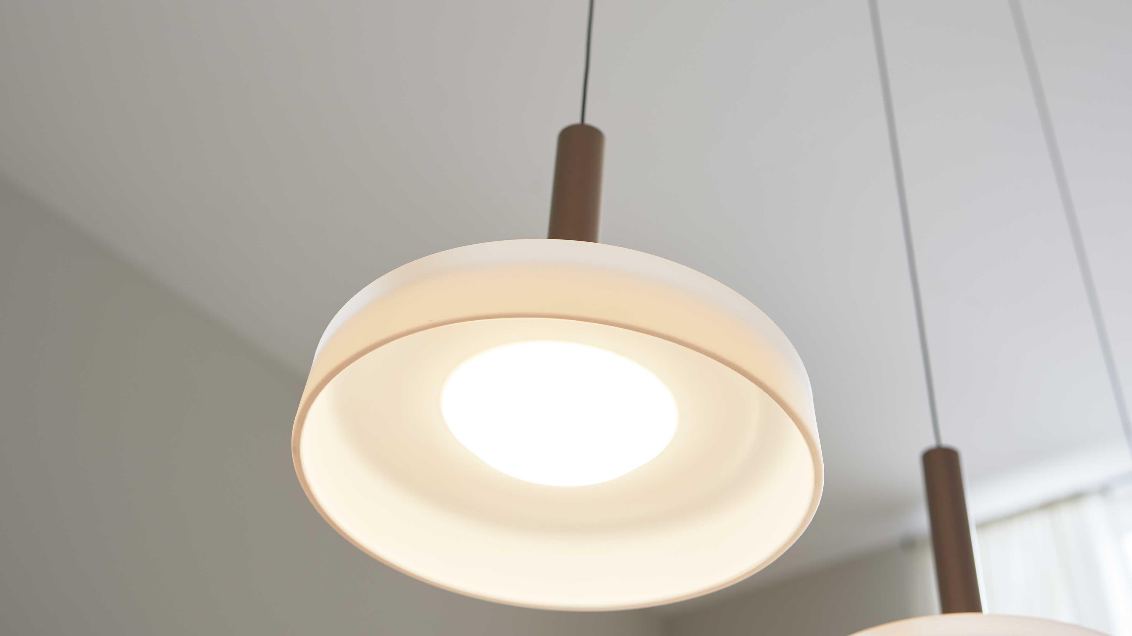 Runde Pendelleuchte mit doppellagigem weißen Schirm und warm leuchtendem Zentrum, zylindrische Aufhängung und zweite Lampe am Bildrand