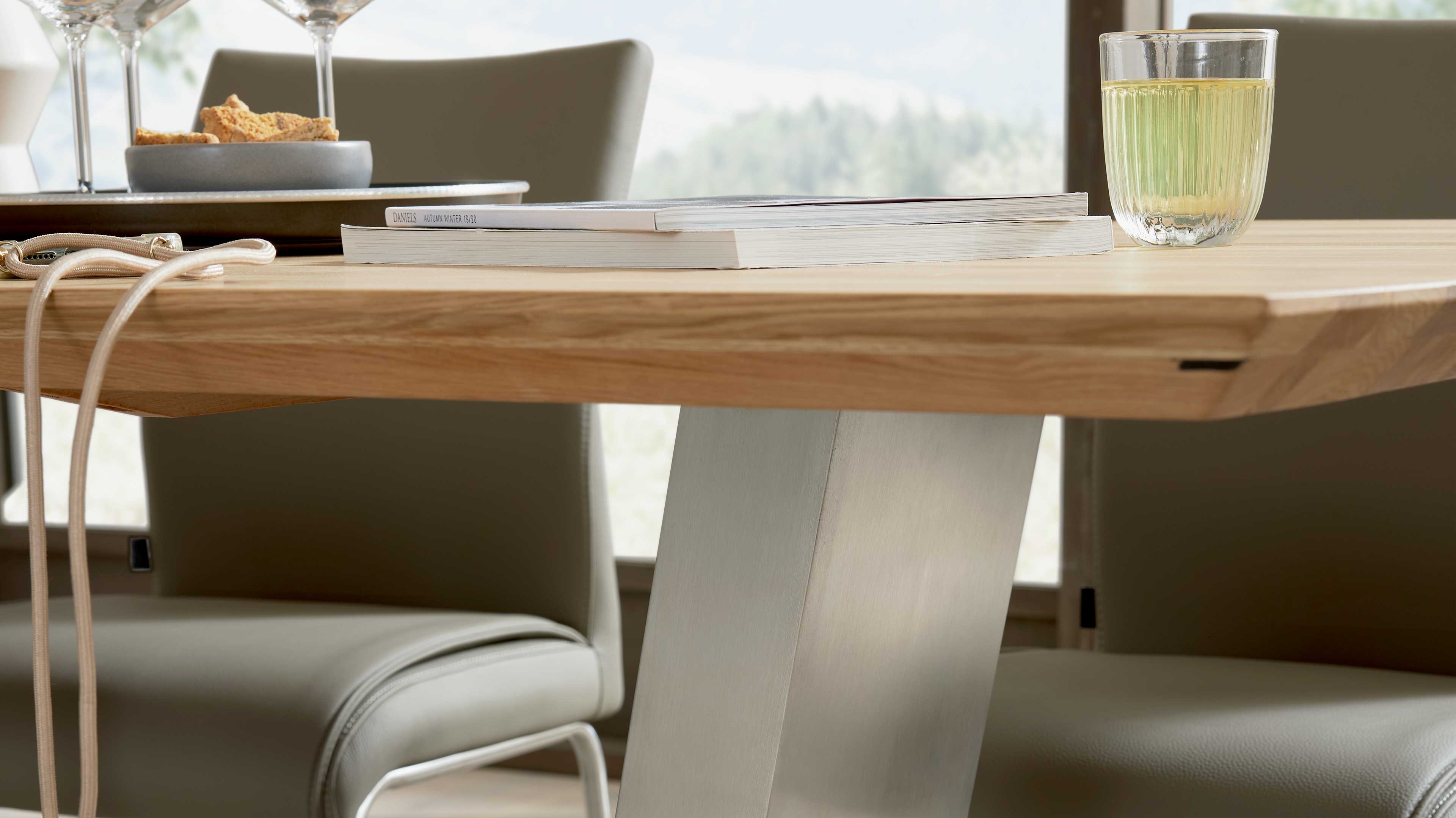 Heller Holztisch mit abgeschrägter Kante und schrägem Metalltischbein, darauf zwei Zeitschriften und ein Glas mit hellem Getränk.