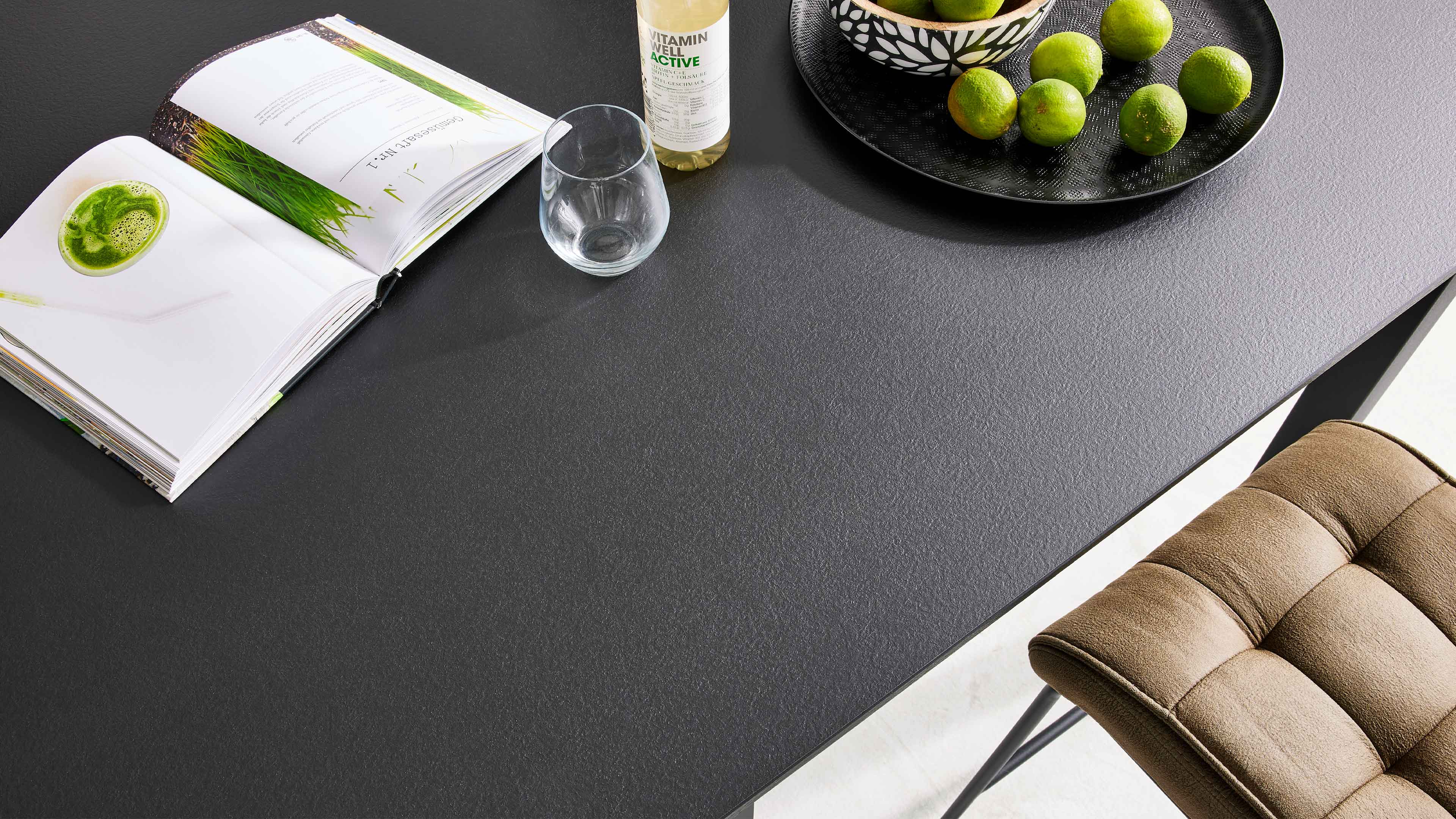 Dunkle, strukturiert wirkende Tischplatte mit offenem Buch, Glasflasche, Trinkglas, schwarzem Tablett mit Limonen und braunem Polsterstuhl.