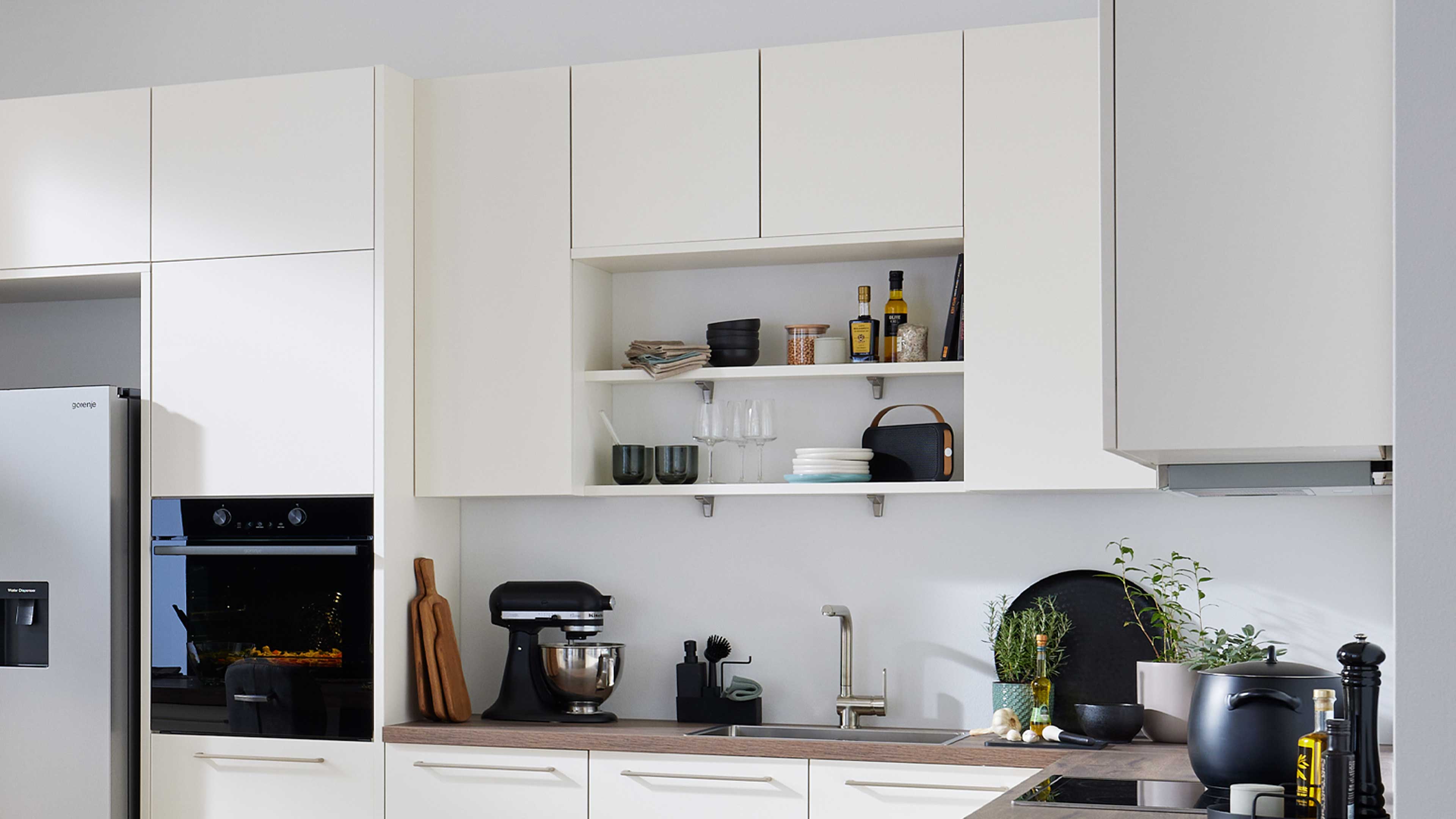 Weiße Oberschränke mit offener Mittelablage über Holzarbeitsplatte mit Spüle, schwarzer Küchenmaschine, Kräutertöpfen und Kochgeschirr.