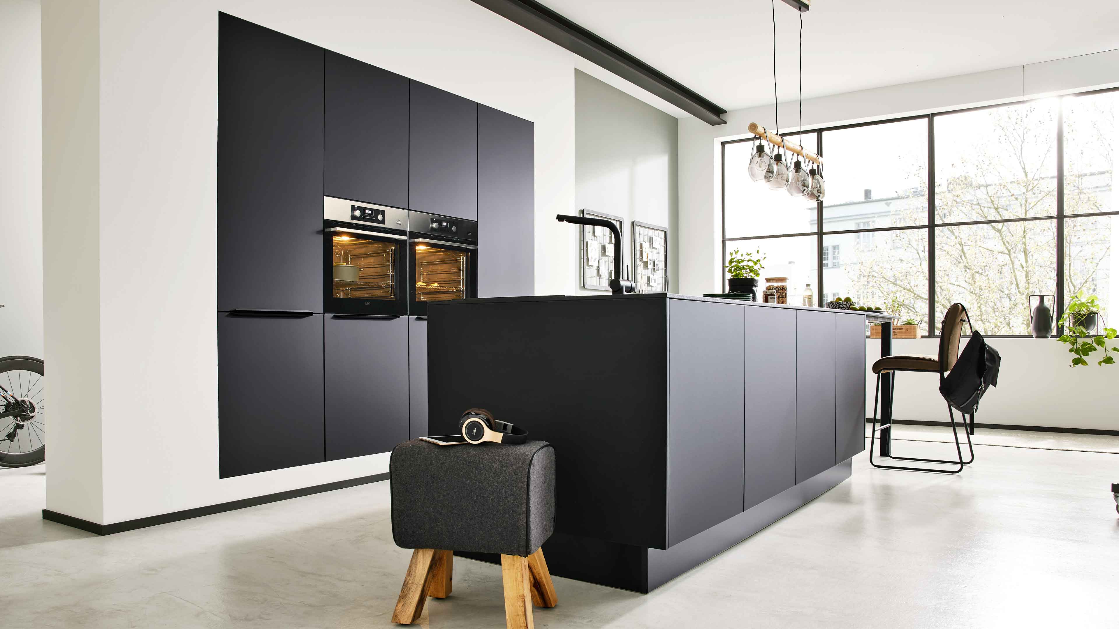 Dunkelgraue Kücheninsel mit Spüle vor hohen Schränken mit zwei Backöfen, davor ein grauer Hocker auf Holzbeinen mit Kopfhörern.