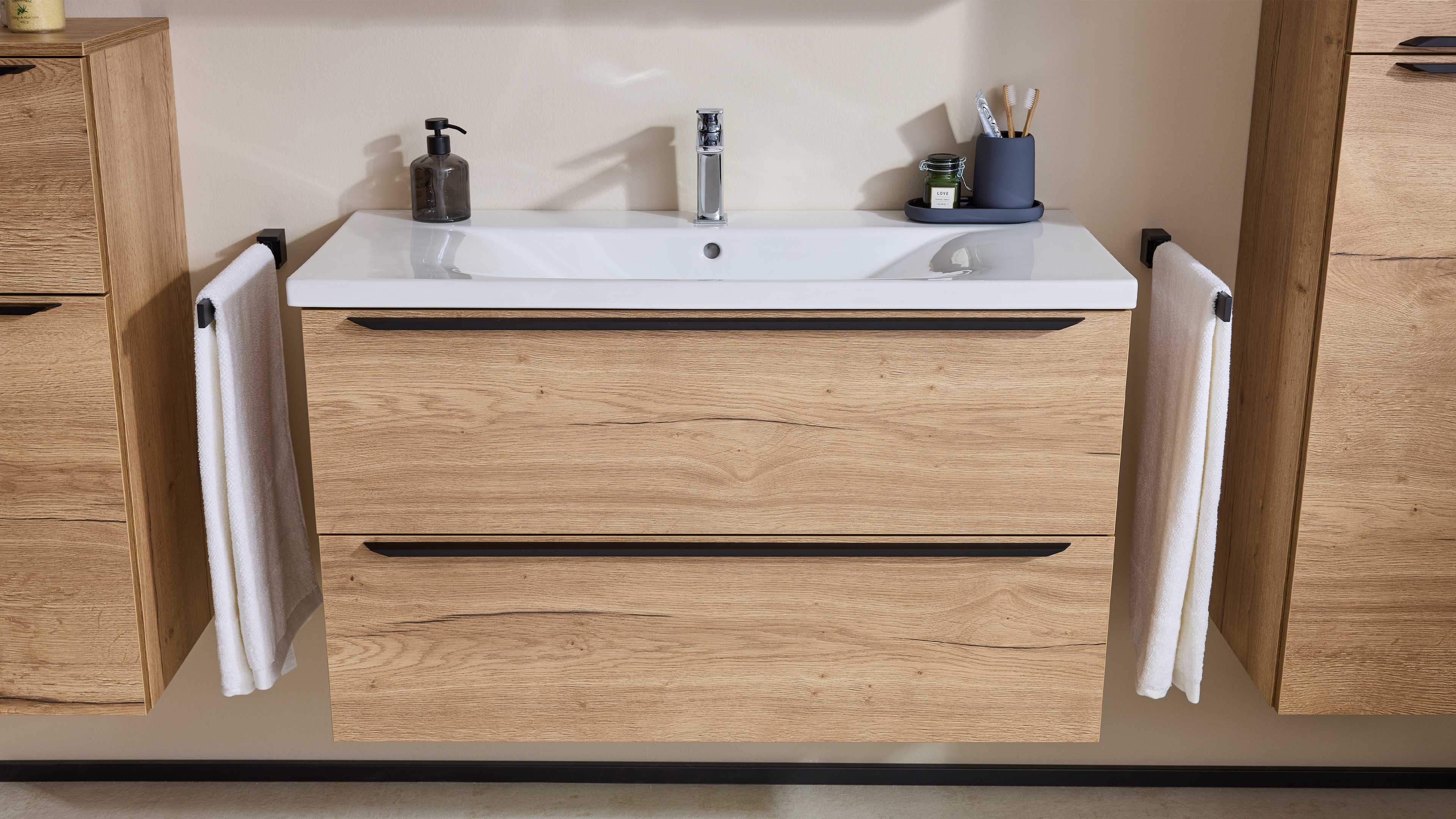 Hängender Holzeffekt-Waschtisch mit weißem Becken, Chromarmatur, links Seifenspender und Handtuch, rechts Zahnputzbecher und Handtuch.