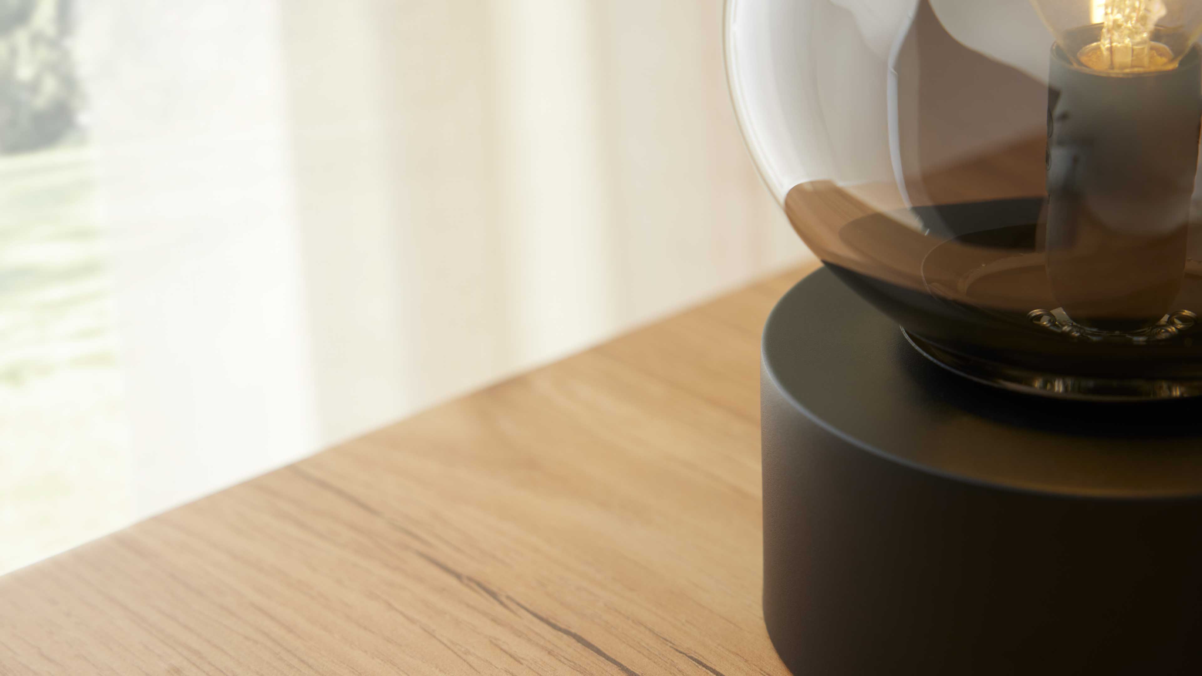 Runder schwarzer Lampensockel mit aufgesetzter Glasglocke und sichtbarer Glühfaden auf hellem Holztisch vor transparenten Vorhängen.