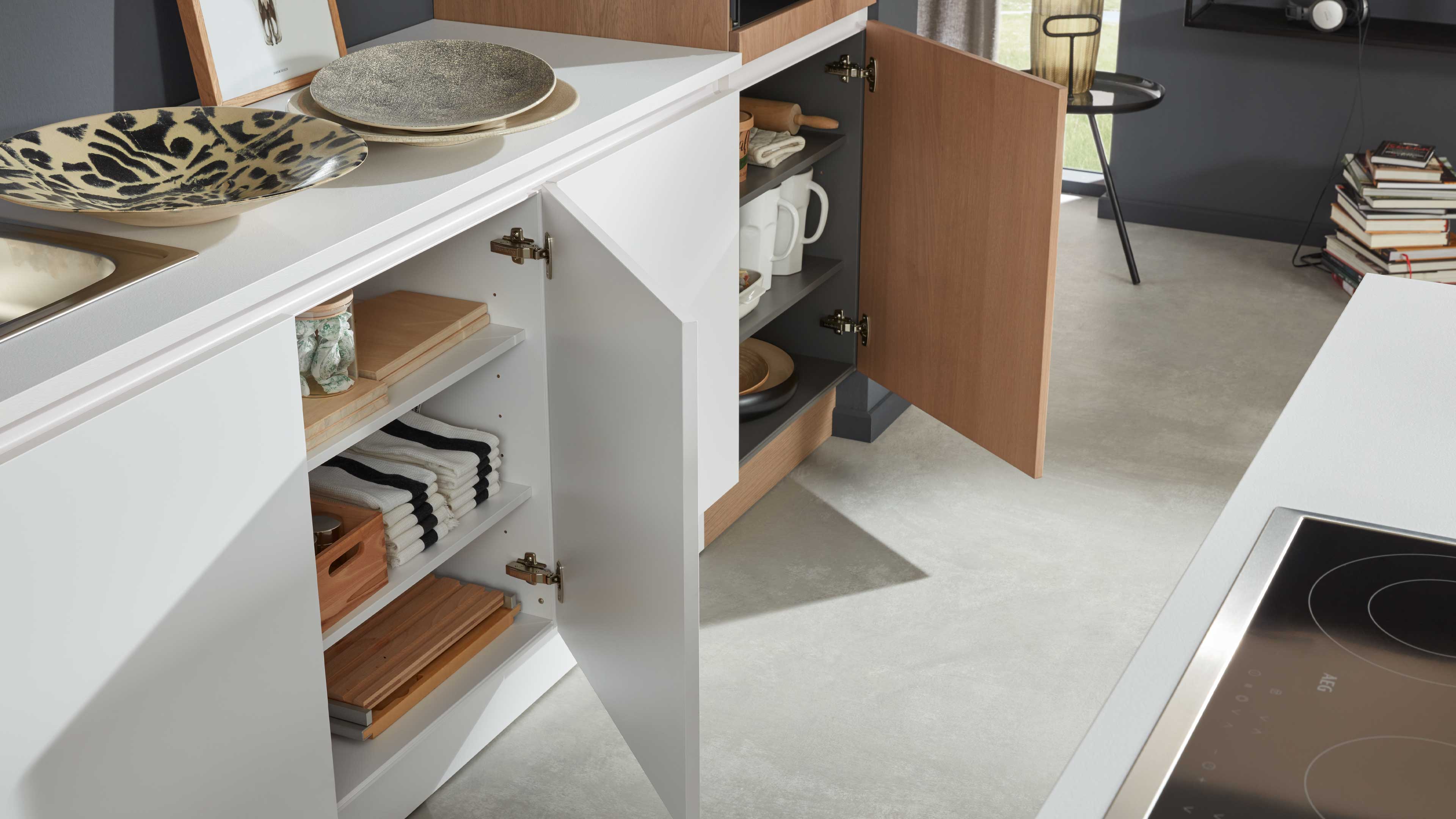 Offene weiße und holzfarbene Küchenunterschränke mit sichtbaren Regalen, gefalteten Handtüchern, Schneidebrettern und weißem Geschirr.