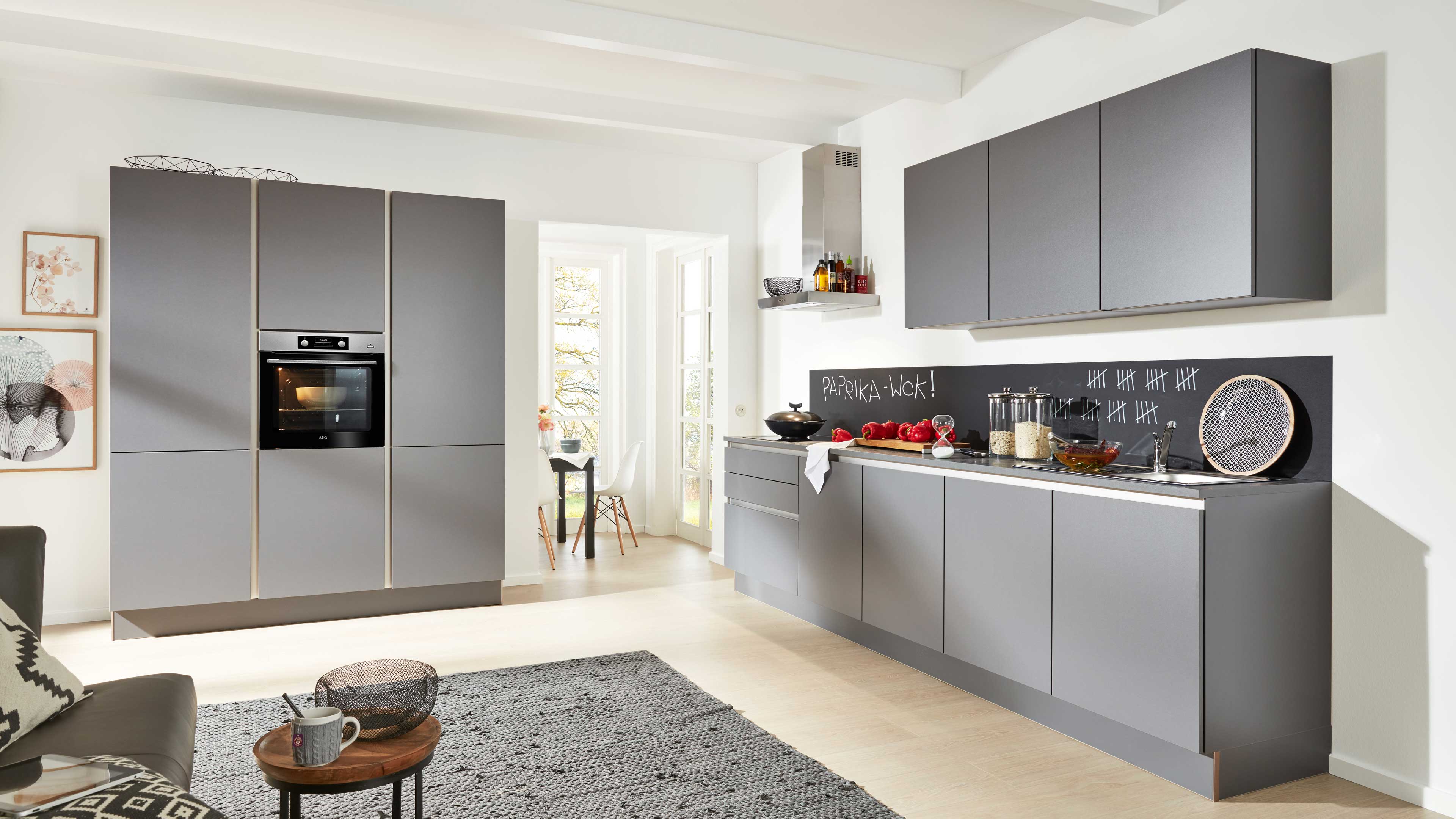 Offene Küche mit mattgrauen, griffarmen Fronten, Hochschrank mit Einbauofen, Hängeschränken und schwarzer Tafelfläche als Spritzschutz.