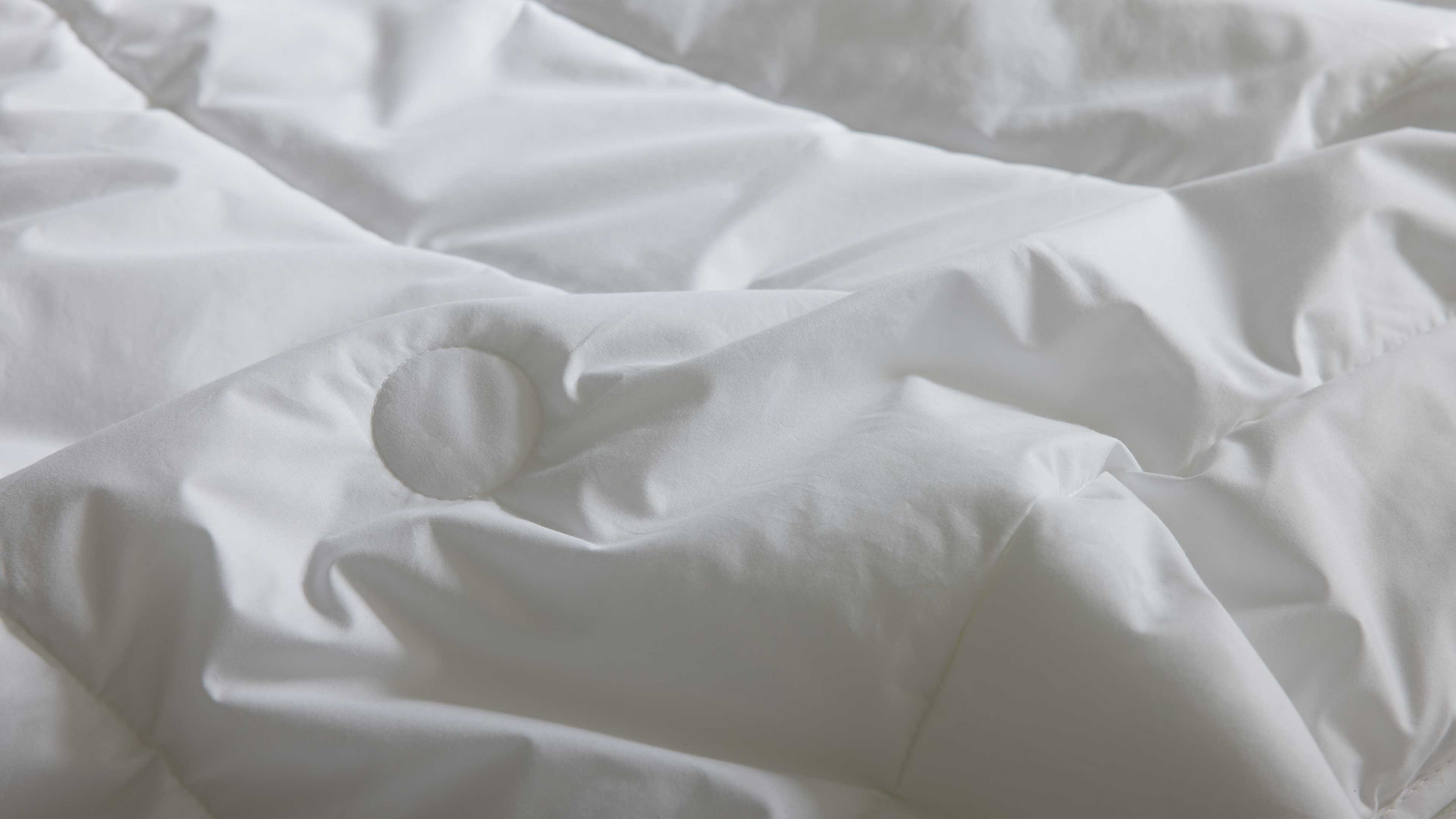 Weiße Bettdecke mit gesteppten Kammern, sichtbarer runder Stegknopf und weiche Falten in der Oberfläche.