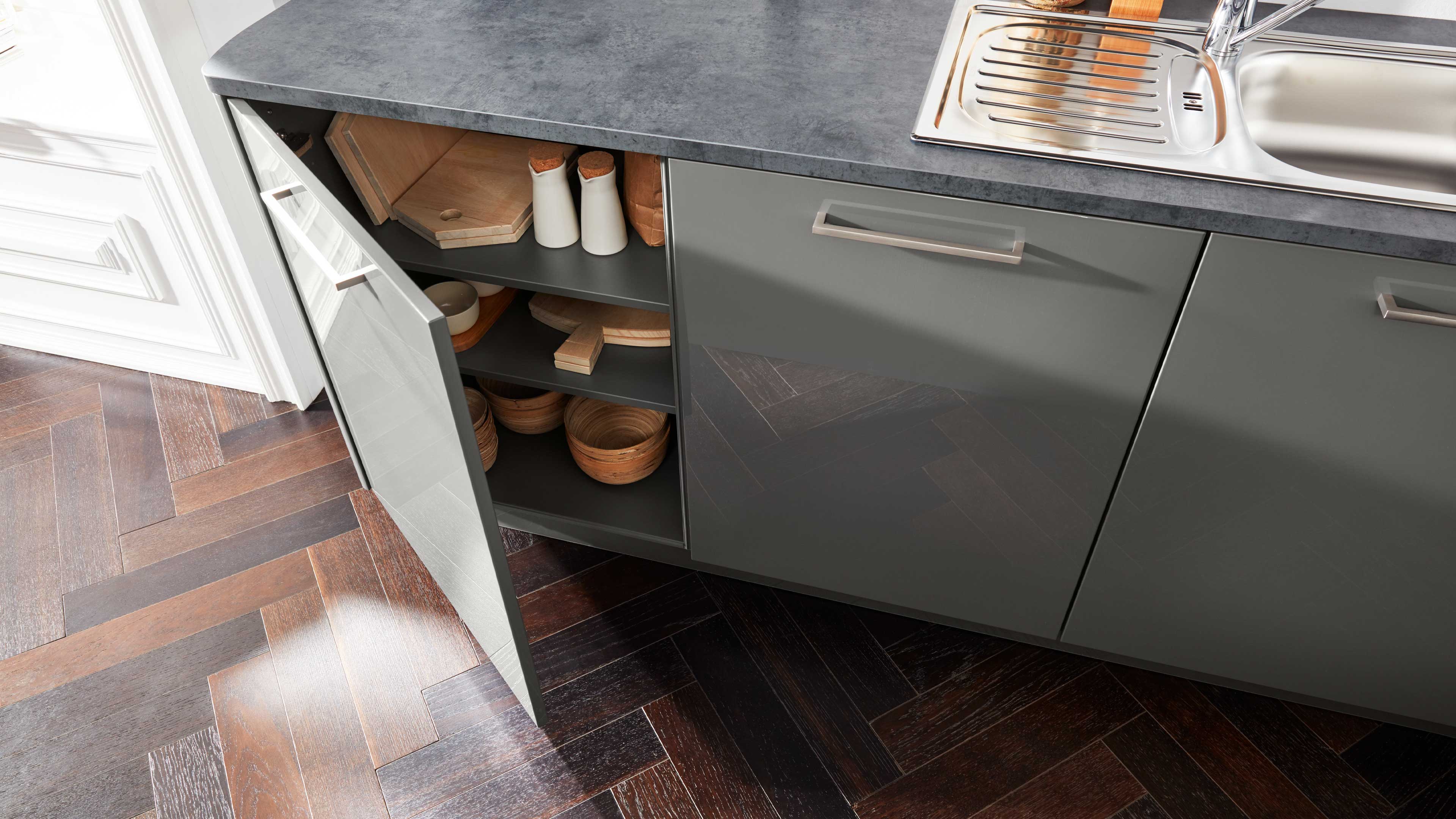 Untere Küchenzeile mit grauen, leicht glänzenden Fronten, grauer Arbeitsplatte, Spüle und offenem Schrank mit Holzschalen und Brettern.