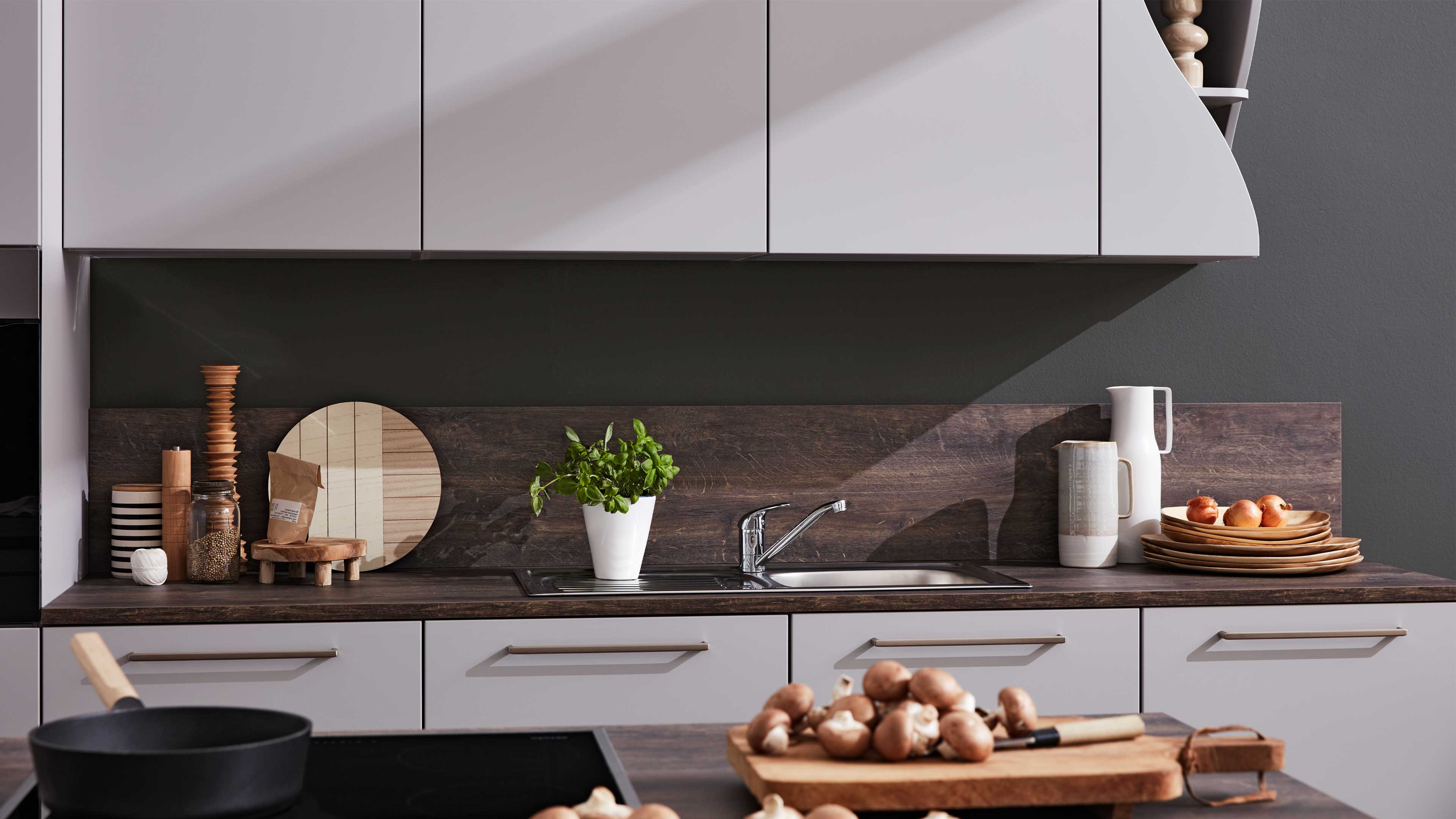 Küchenarbeitsplatte mit eingebauter Edelstahlspüle und Armatur, ein Topf mit Basilikum, rechts Tellerstapel und Krüge, links Gewürzdosen.