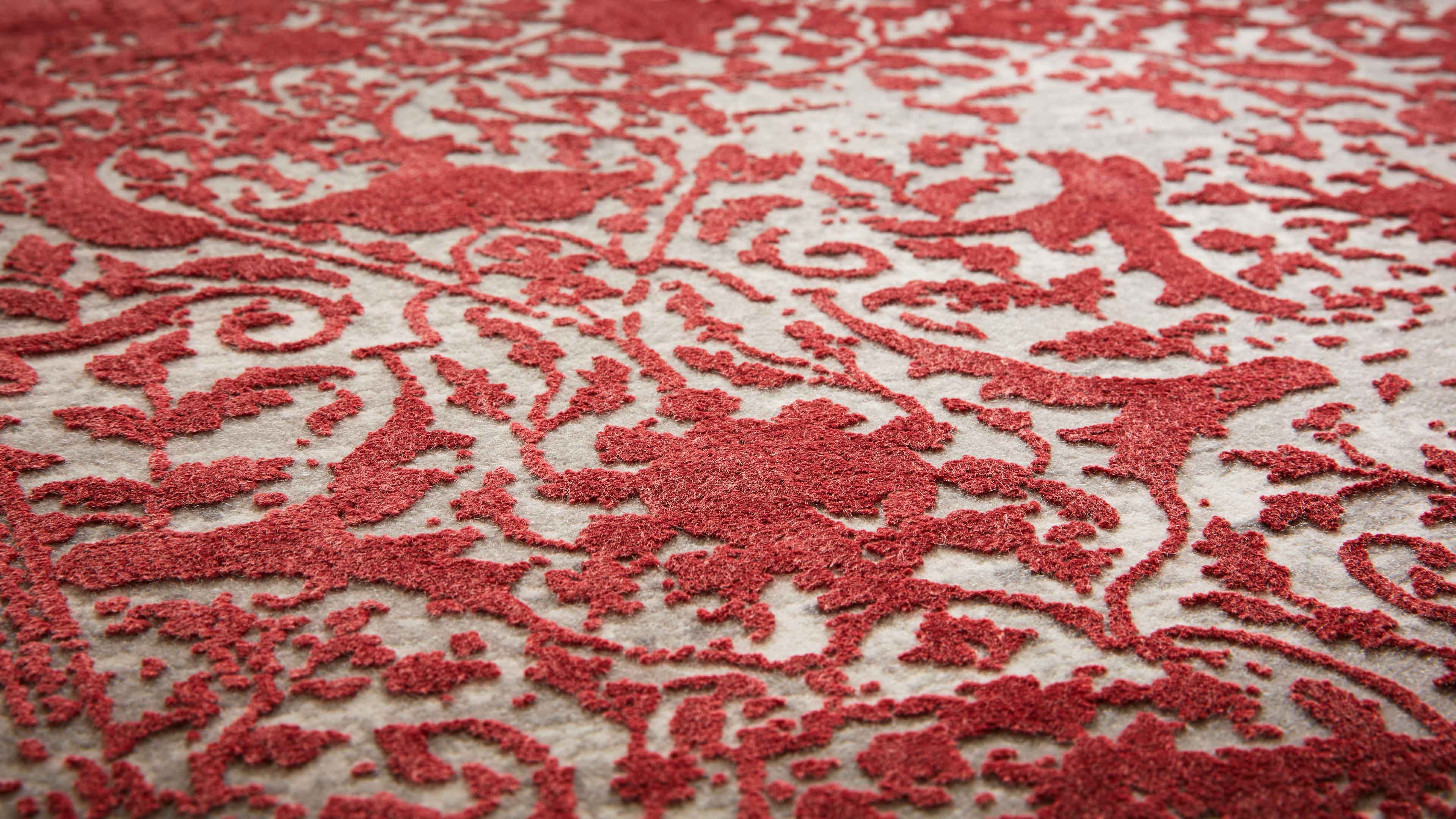 Nahaufnahme eines Teppichs mit roten, erhabenen Blumen- und Rankenmustern auf hellbeigem, strukturiertem Hintergrund.