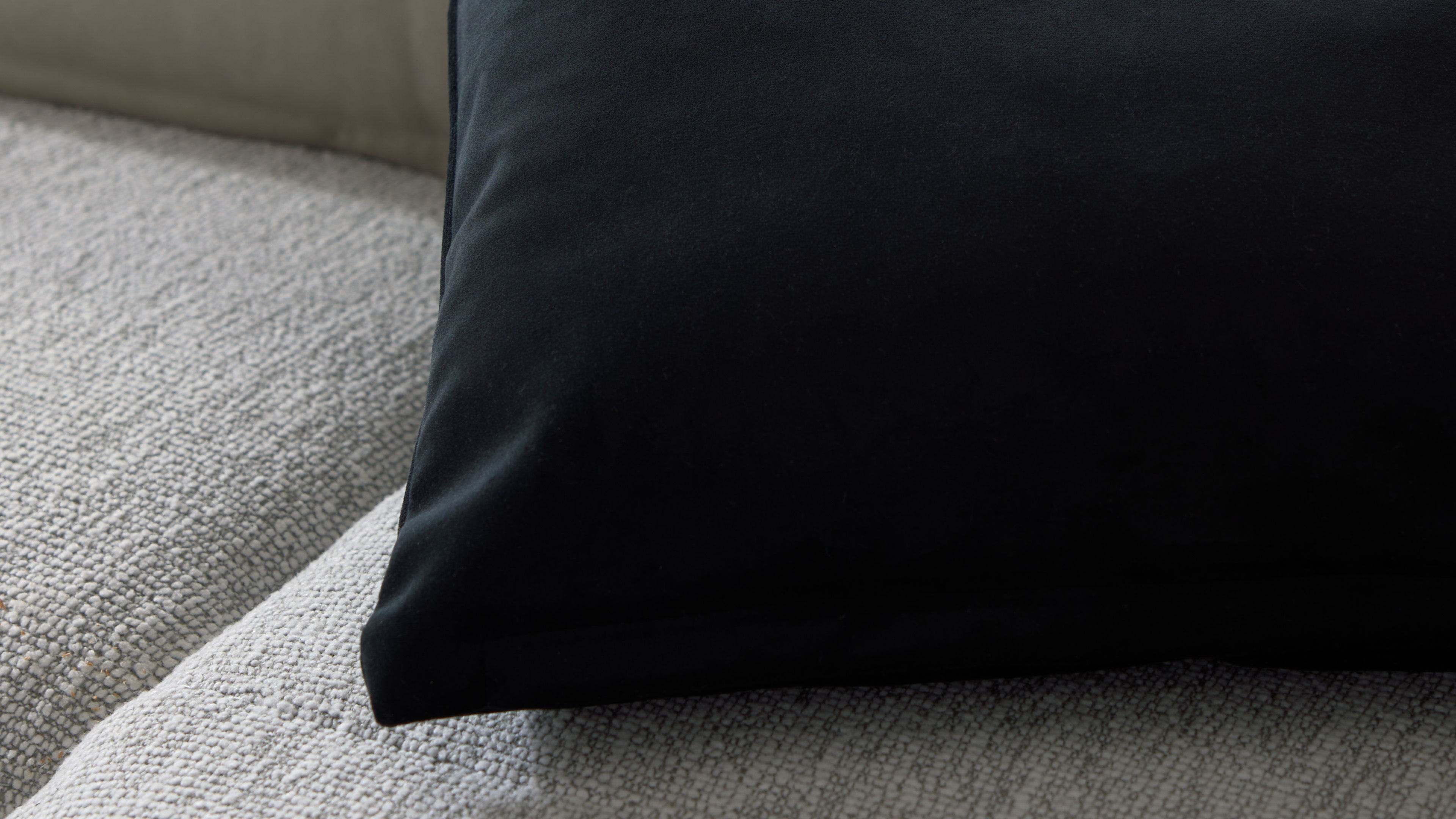Schwarzes Kissen liegt auf hellgrauem, grob gewebtem Polsterbezug; untere Kissenecke ist sichtbar.