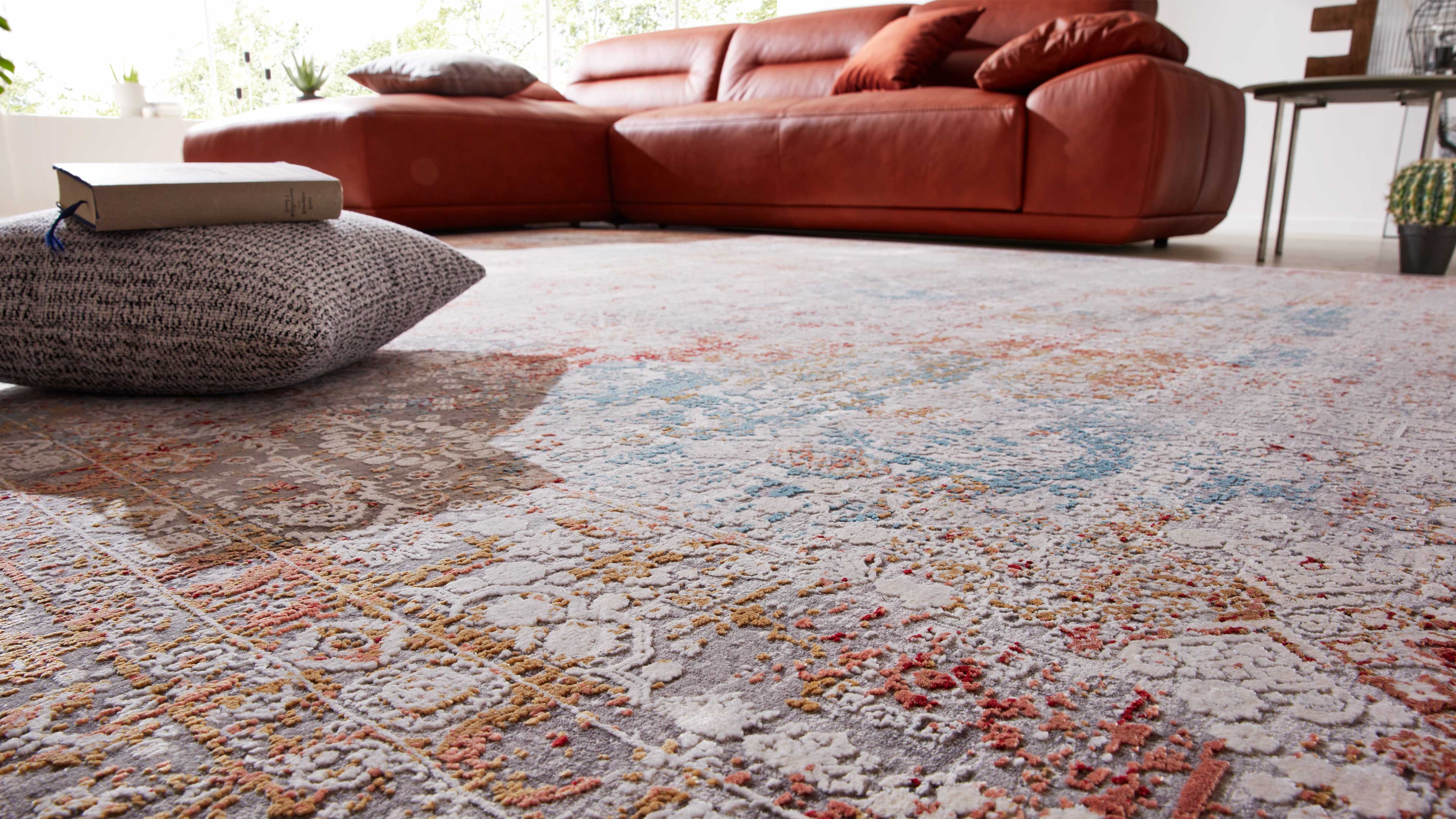 Großflächiger, farbig gemusterter Wohnzimmerteppich im Vordergrund, dahinter rotes Leder-Ecksofa, graues Bodenkissen mit Buch.