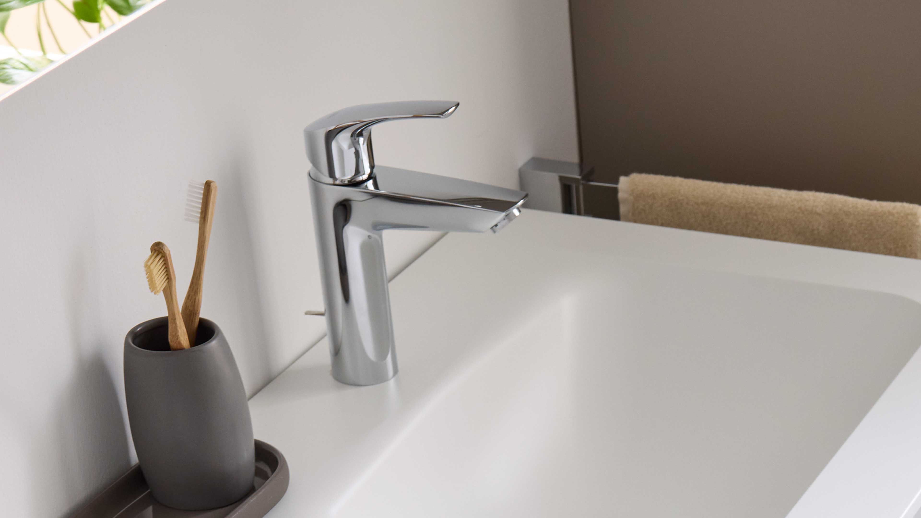 Chromfarbene Einhebelarmatur über weißer Keramikspüle, links ein grauer Becher mit zwei Bambuszahnbürsten, rechts ein beiges Handtuch.