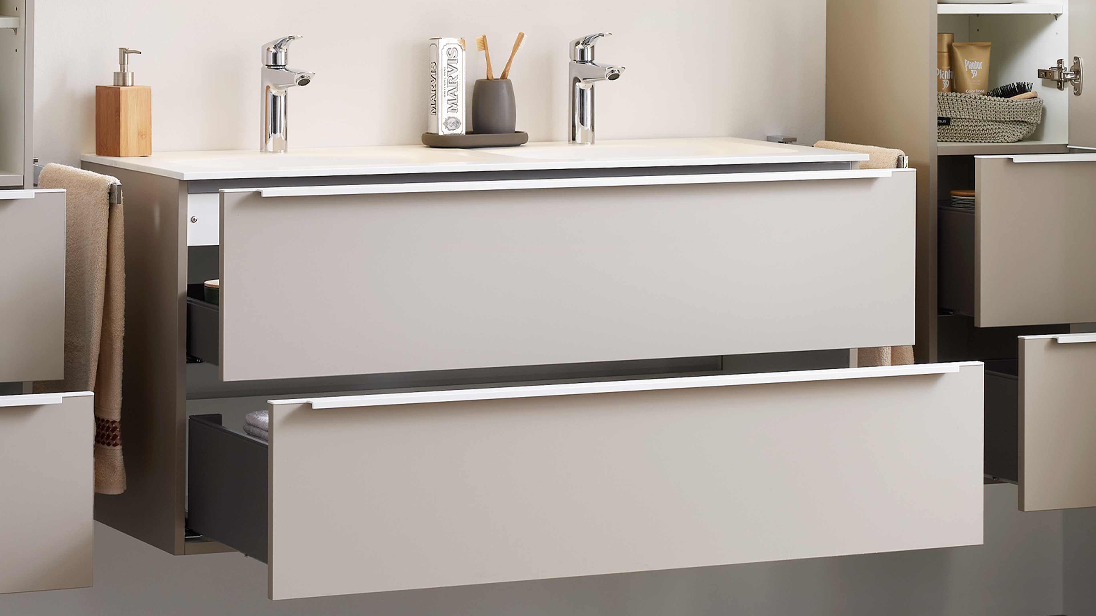 Wandhängender Waschplatz mit zwei Chromarmaturen auf weißer Platte, zwei großen Schubladen und offenem Seitenschrank mit Handtuch.