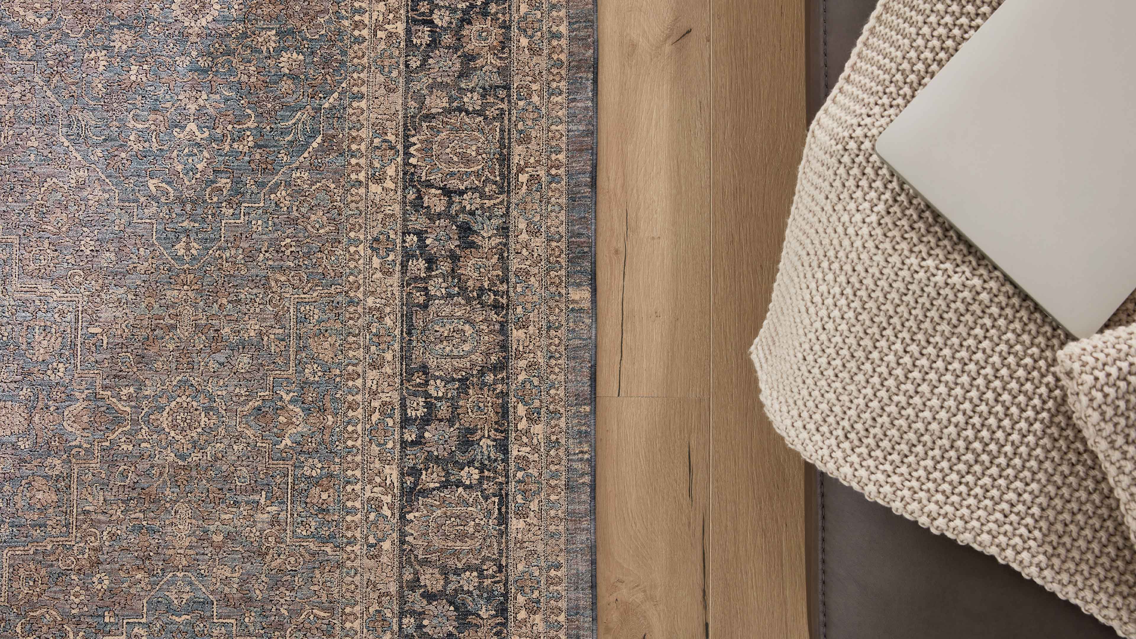 Orientalisch gemusterter Teppich links, rechts sichtbare helle Holzdielen und ein beigefarbener Stricküberwurf auf dem Sofa mit Laptop.