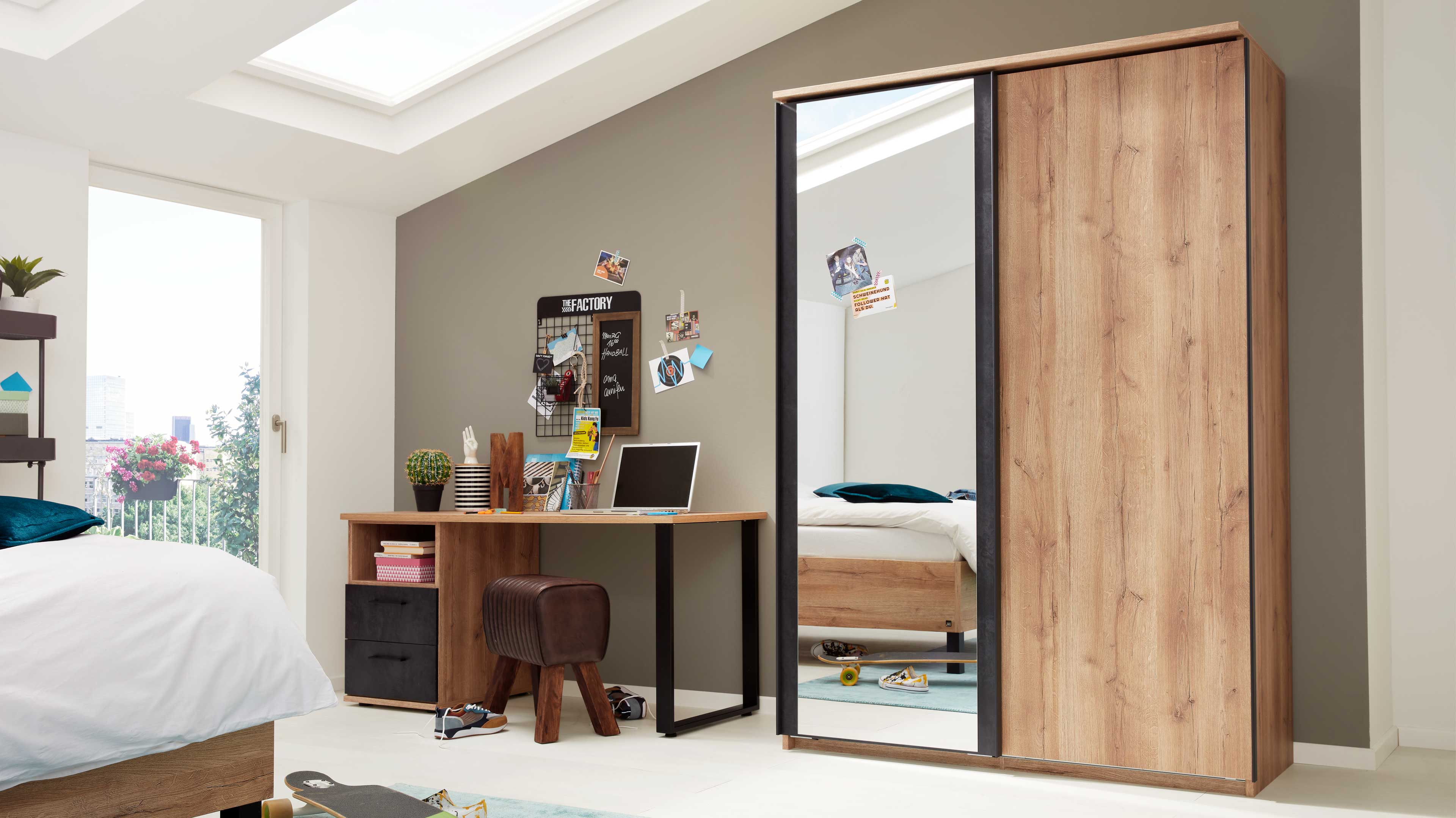 Großer Holzkleiderschrank mit einer Spiegeltür und einer Holz-Schiebetür, daneben Schreibtisch mit Laptop und Hocker.