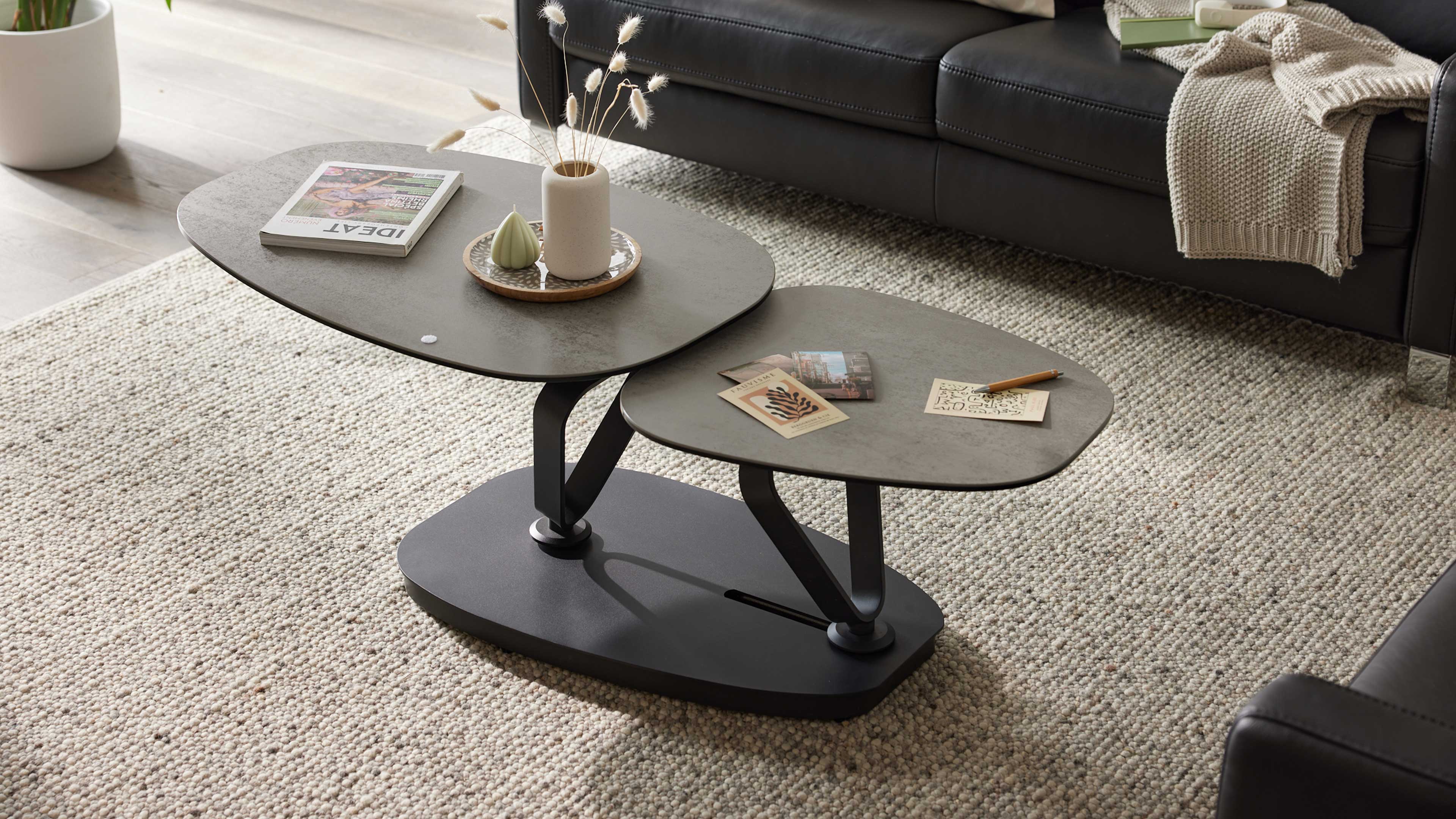 Zwei ovale, graue Couchtische auf schwarzer Basis auf hellem Grobstrick-Teppich, mit Vase, Kerze, Zeitschrift und Sofa im Hintergrund.