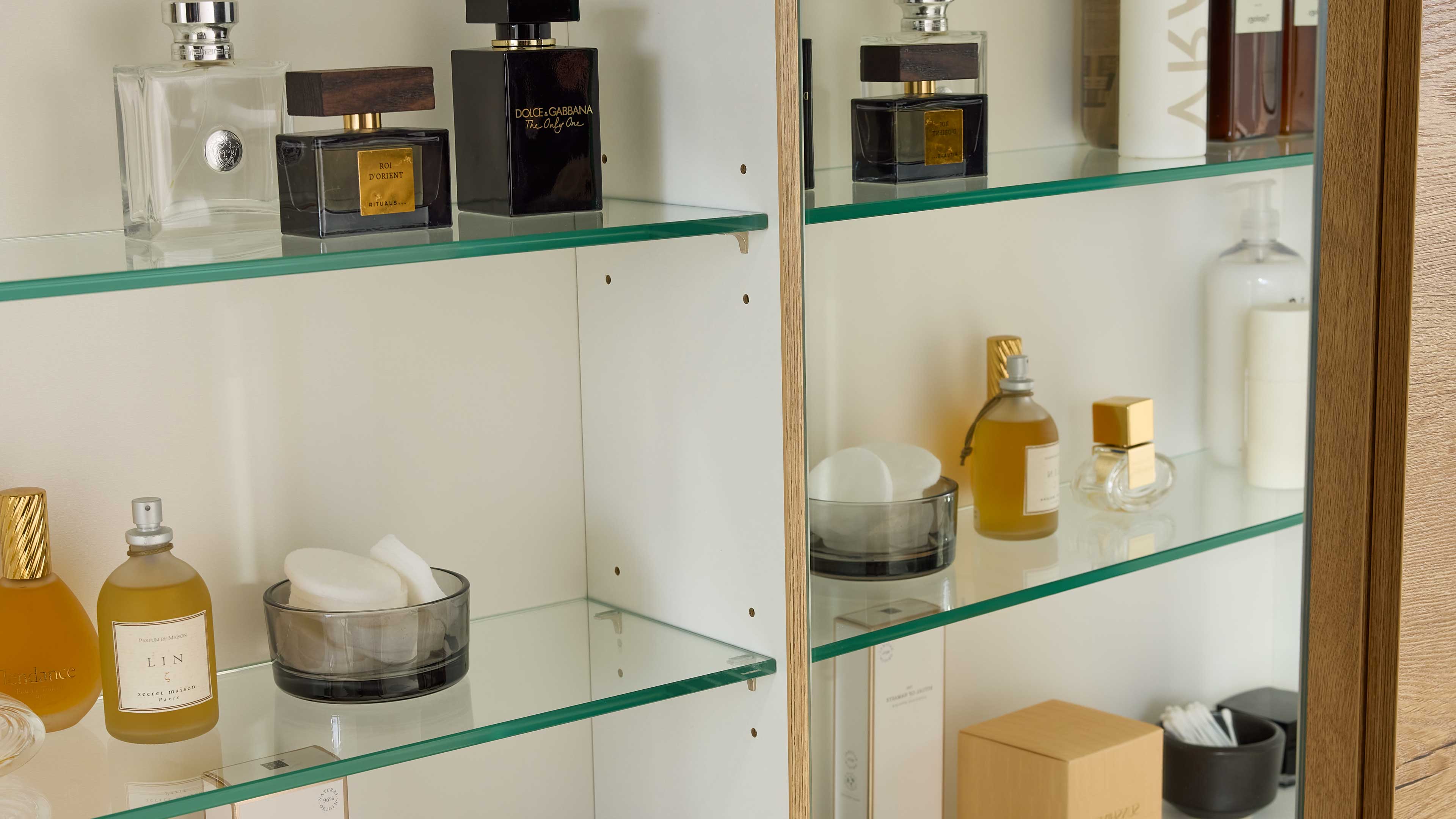Holzgerahmter Badschrank mit weißen Innenwänden und Glasregalen, bestückt mit Parfüms, Glasschale mit Wattepads und Pflegeflaschen.