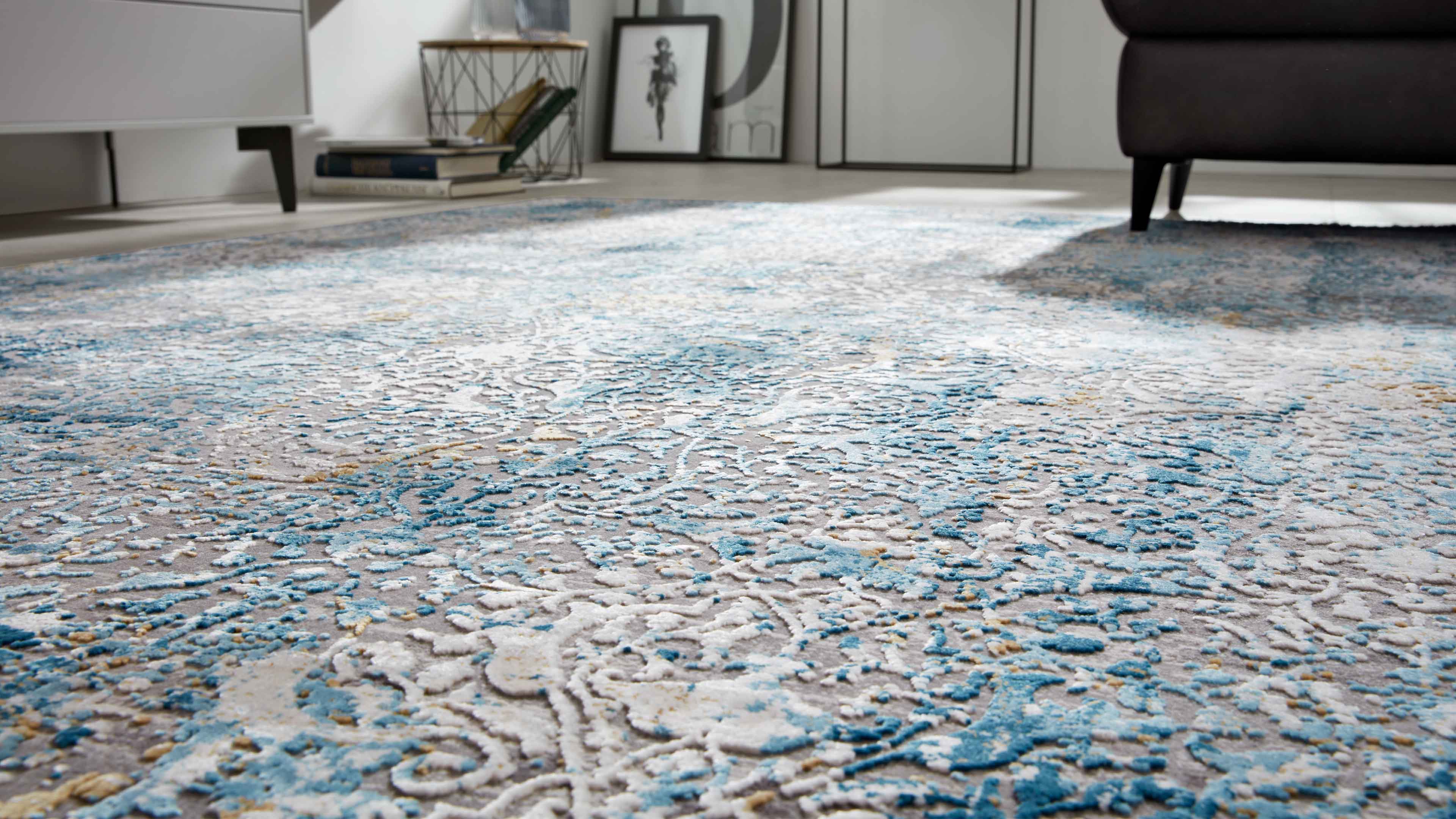 Teppich mit erhabenem floralem Reliefmuster in Blau, Weiß und Beige; rechts Sofa‑bein, links Sideboardbein und Bücher im Hintergrund.