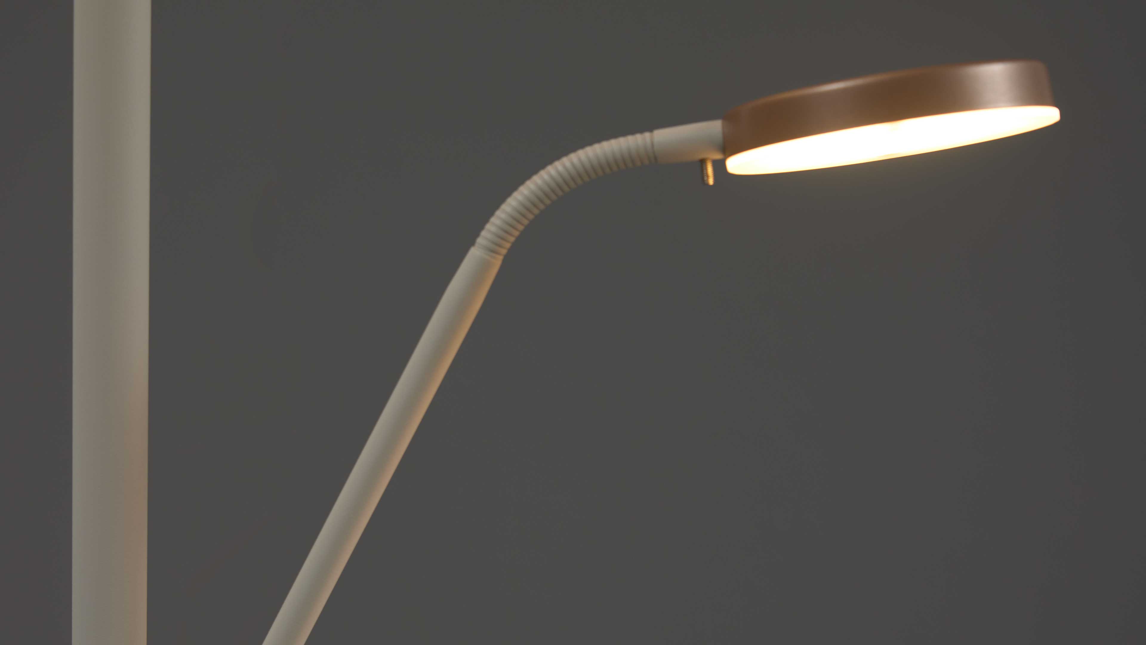 Schwenkbare Leselampe mit dünnem weißem Stab, geriffeltem Gelenk und rundem, warm beleuchtetem Lampenschirm vor dunklem Hintergrund.