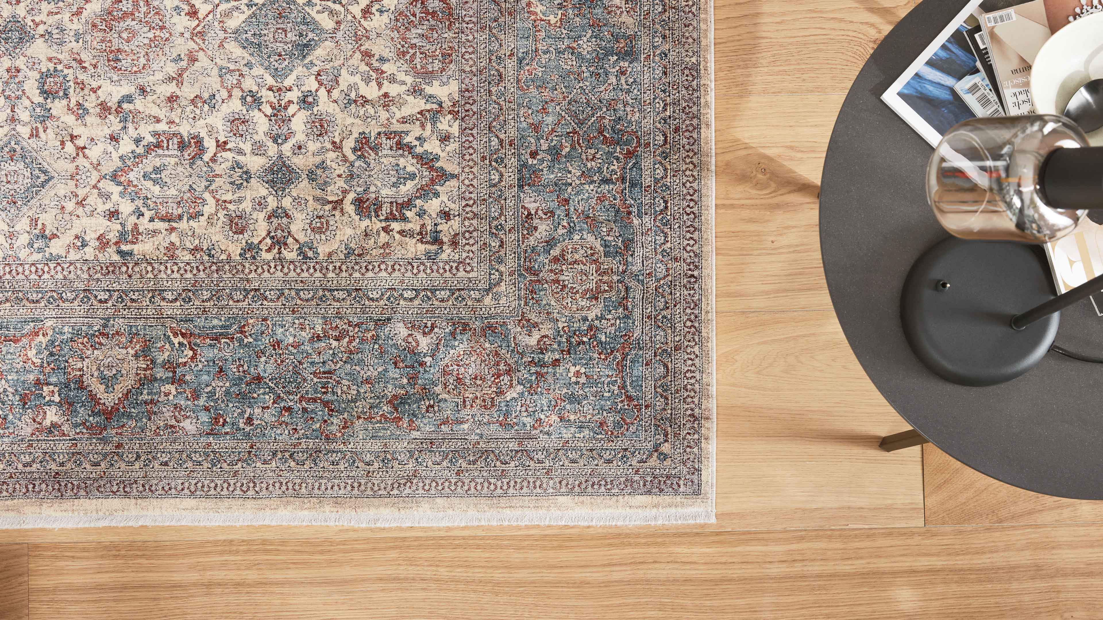 Orientalisch gemusterter Teppich in Beige, Blau und Rost auf hellem Holzboden, rechts ein runder Beistelltisch mit schwarzer Lampe.
