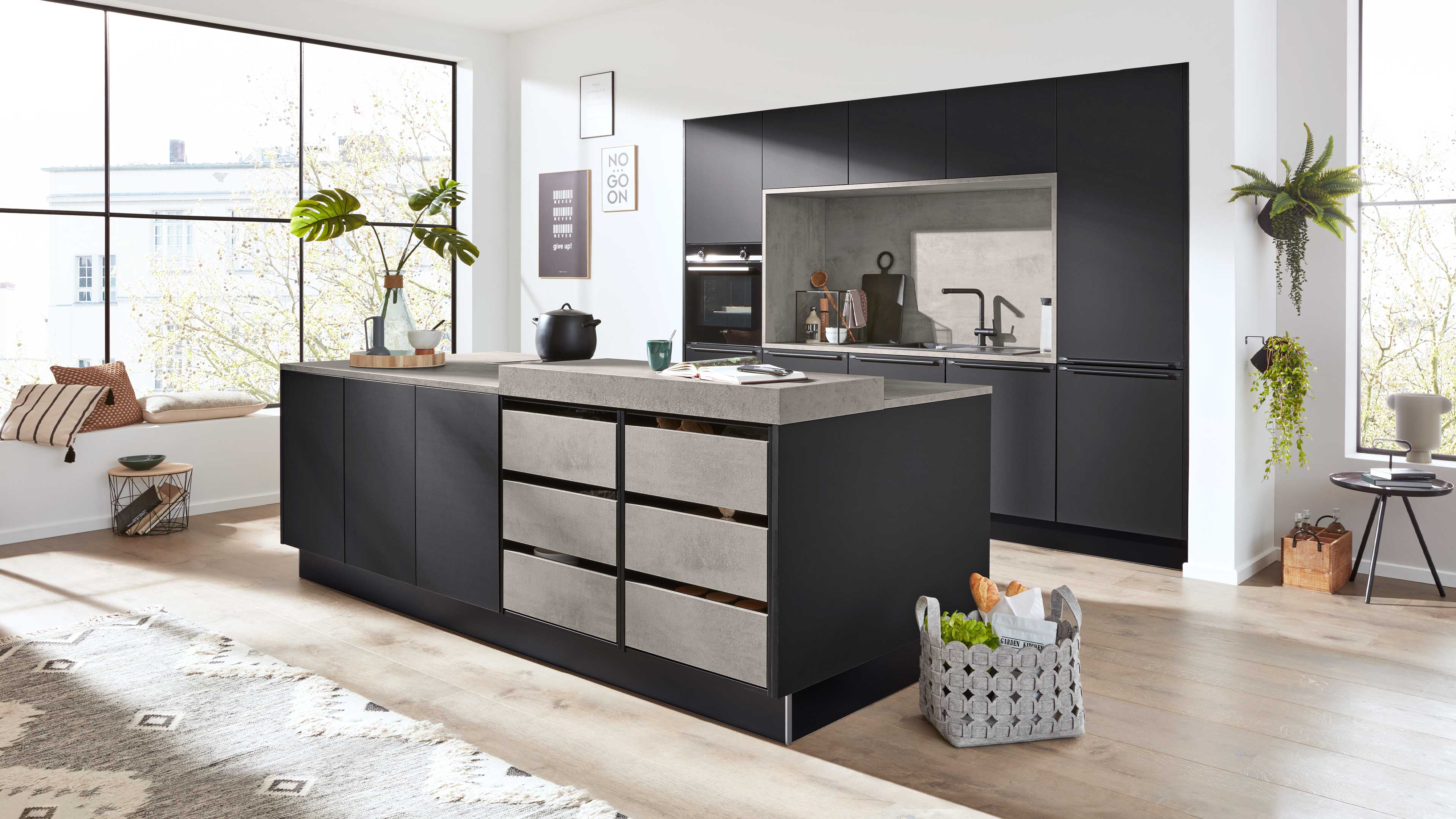 Kücheninsel mit dunkelgrauen Schränken und hellgrauen Schubladen, im Hintergrund Wandküche, große Fenster, Pflanzen und Korb mit Brot.