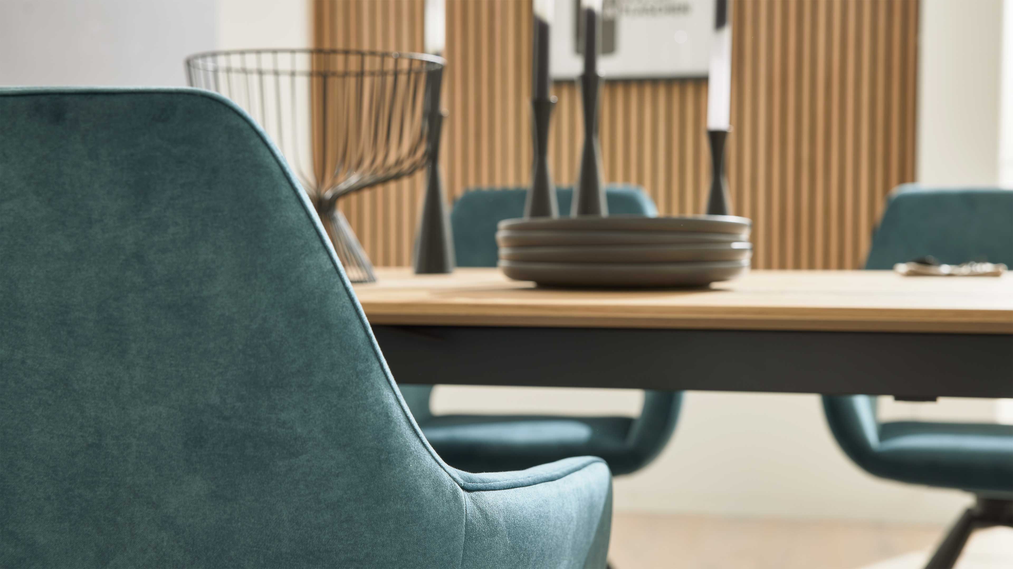 Türkisfarbener Polsterstuhl im Vordergrund, dahinter Holztisch mit grauen Tellern, schwarzen Kerzenhaltern und Drahtkorb vor Lamellenwand.