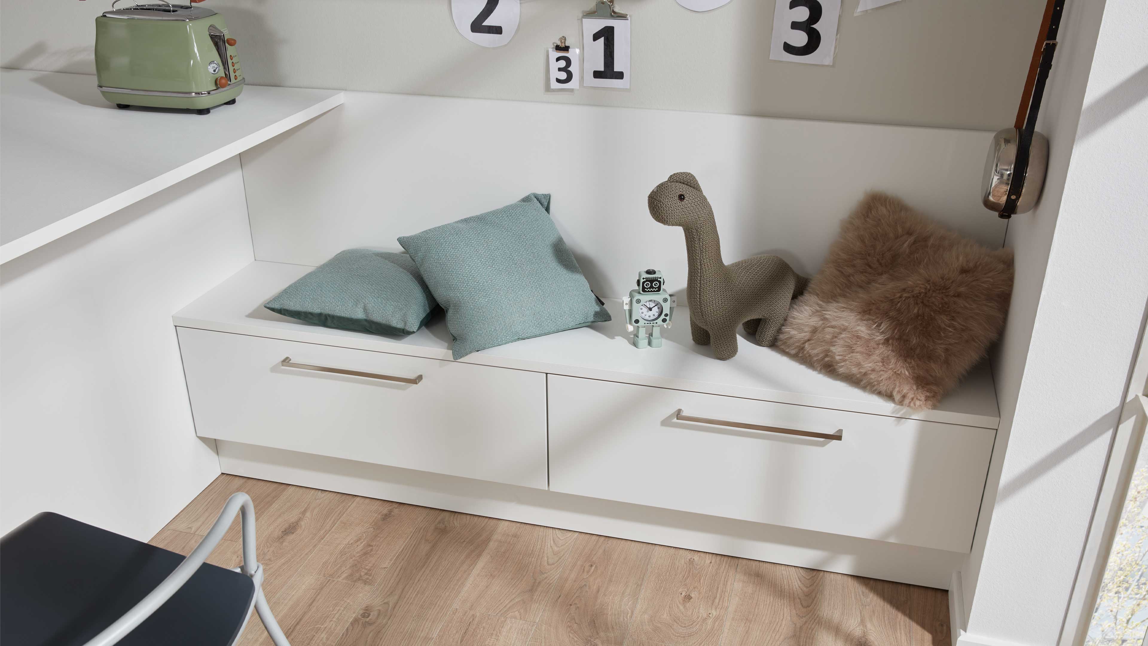 Weiße eingebaute Sitzbank mit zwei Schubkästen, silbernen Griffen, mintgrünen Kissen, gestricktem Dinosaurier und braunem Fellkissen.