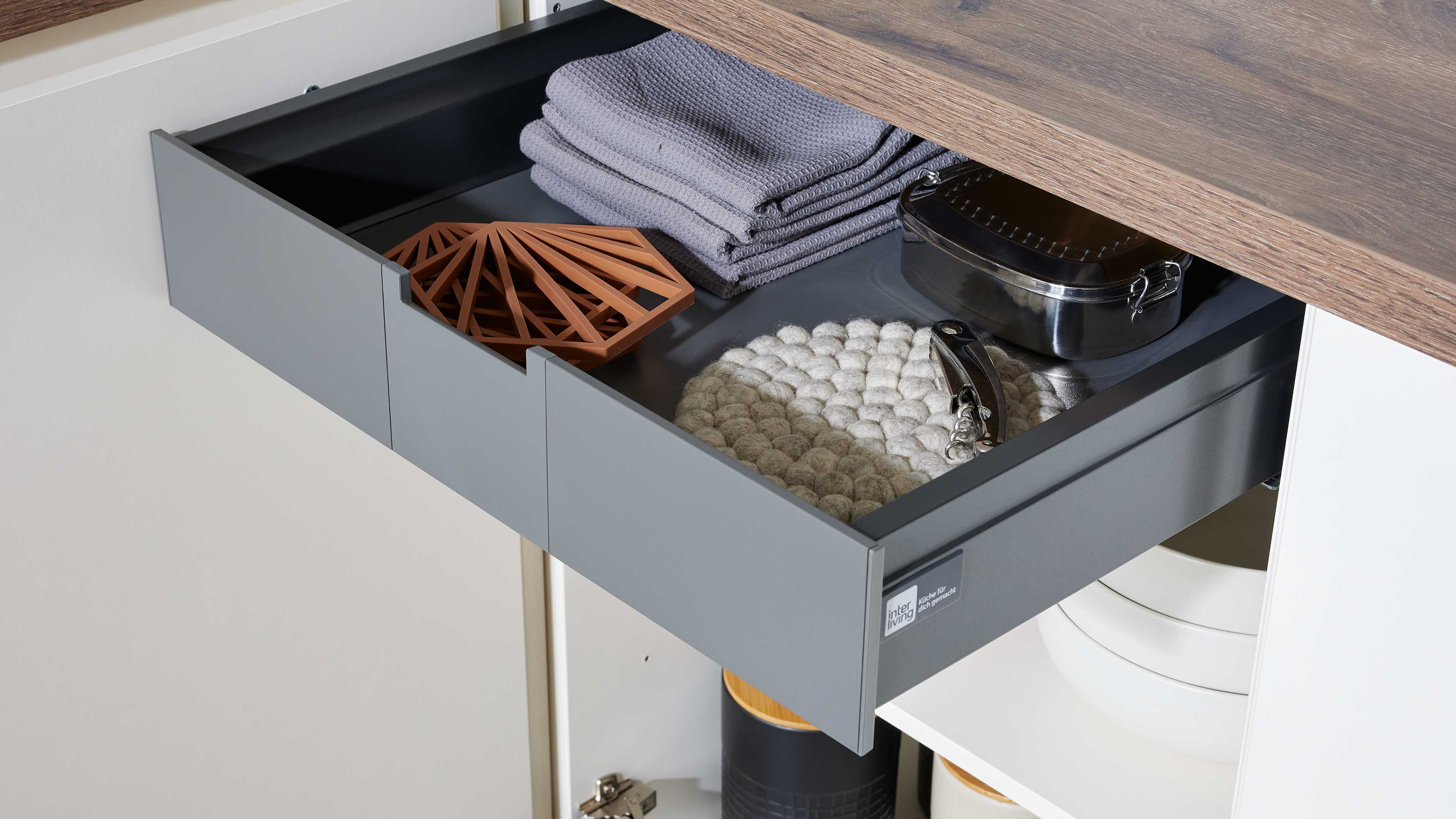 Geöffnete graue Schublade mit gefalteten grauen Tüchern, terrakottafarbenem Untersetzer, runder Filzmatte, Edelstahlbox und Dosenöffner.
