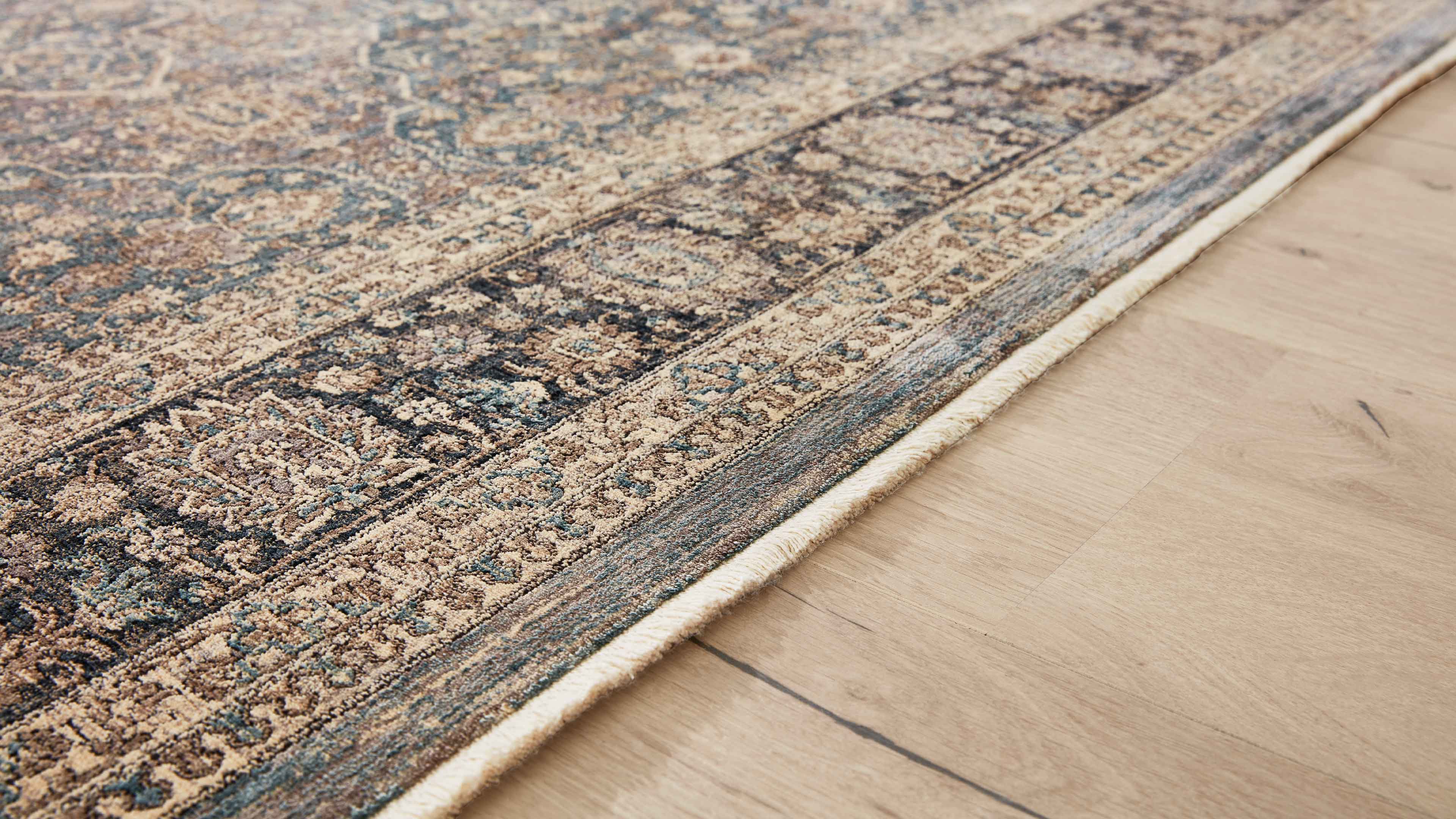 Orientteppich mit floraler Bordüre in Beige, Blau und Dunkelbraun, schräg auf hellem Holzfußboden liegend.