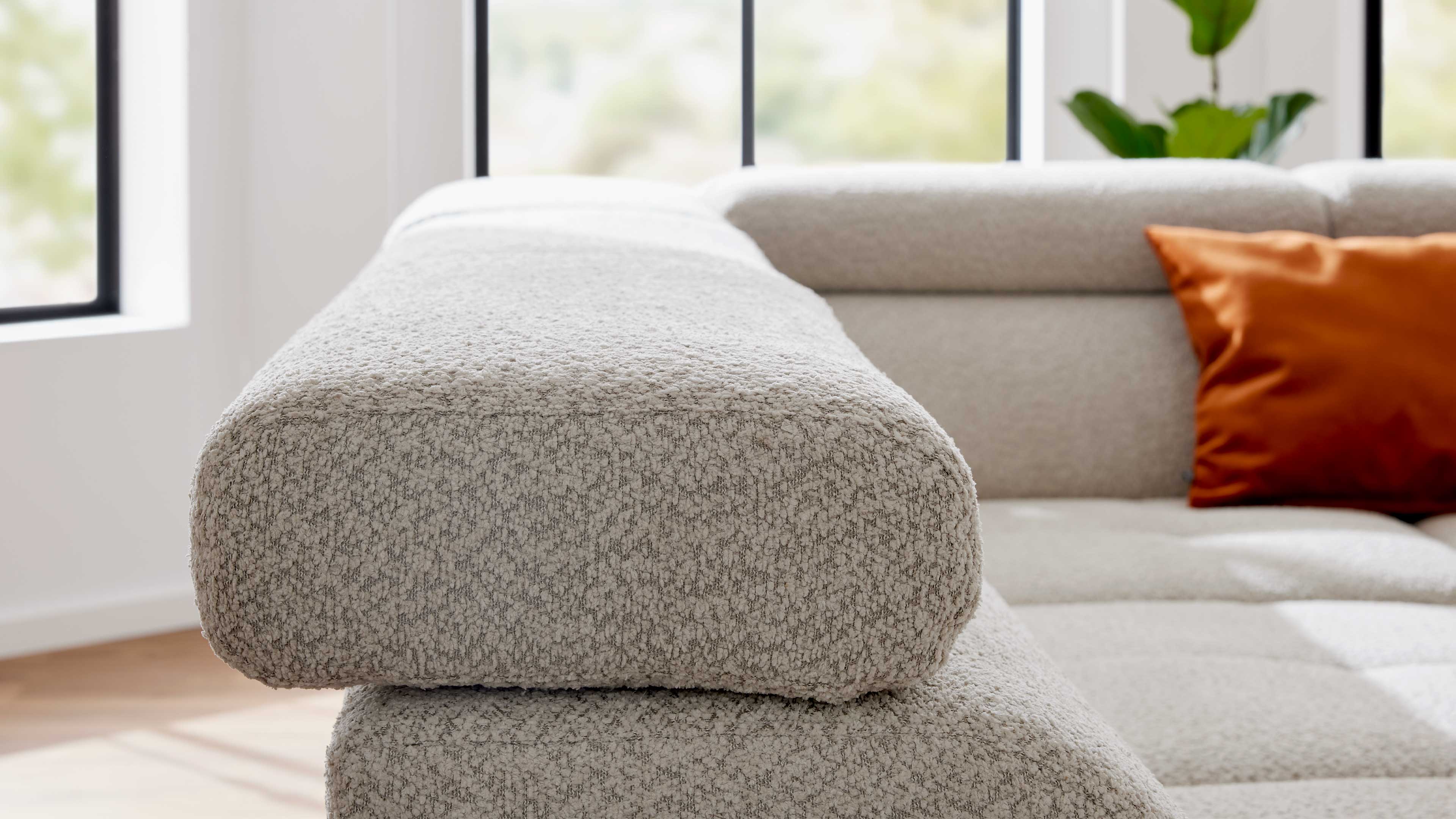 Nahaufnahme einer hellgrauen, abgerundeten Sofaarmlehne mit strukturierter Polsterung, im Hintergrund ein orangefarbenes Kissen und Fenster.