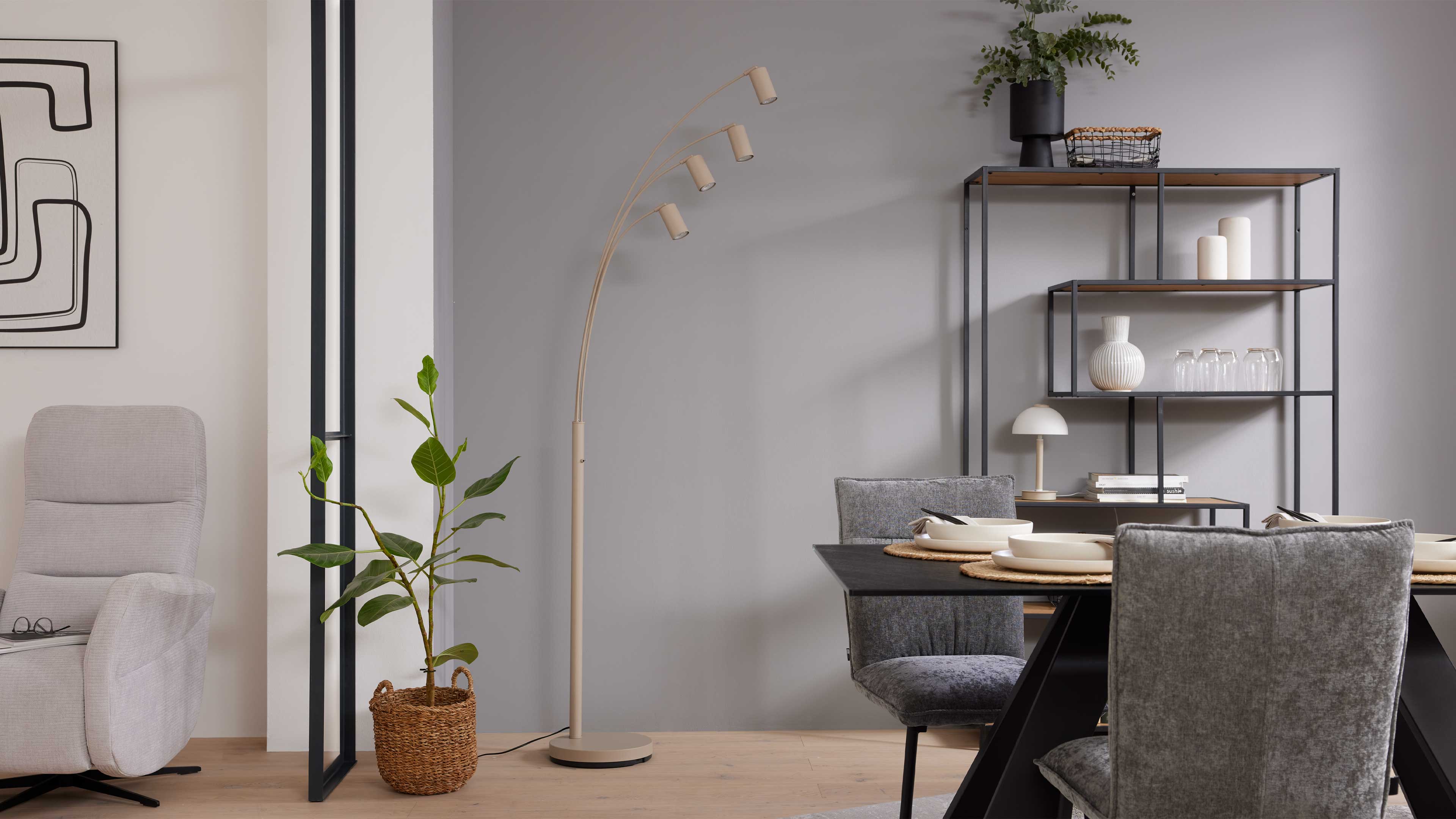 Beige, gebogene Stehleuchte mit vier zylinderförmigen Leuchtenköpfen neben einem Korb mit Zimmerpflanze, Esstisch und offenem Metallregal.