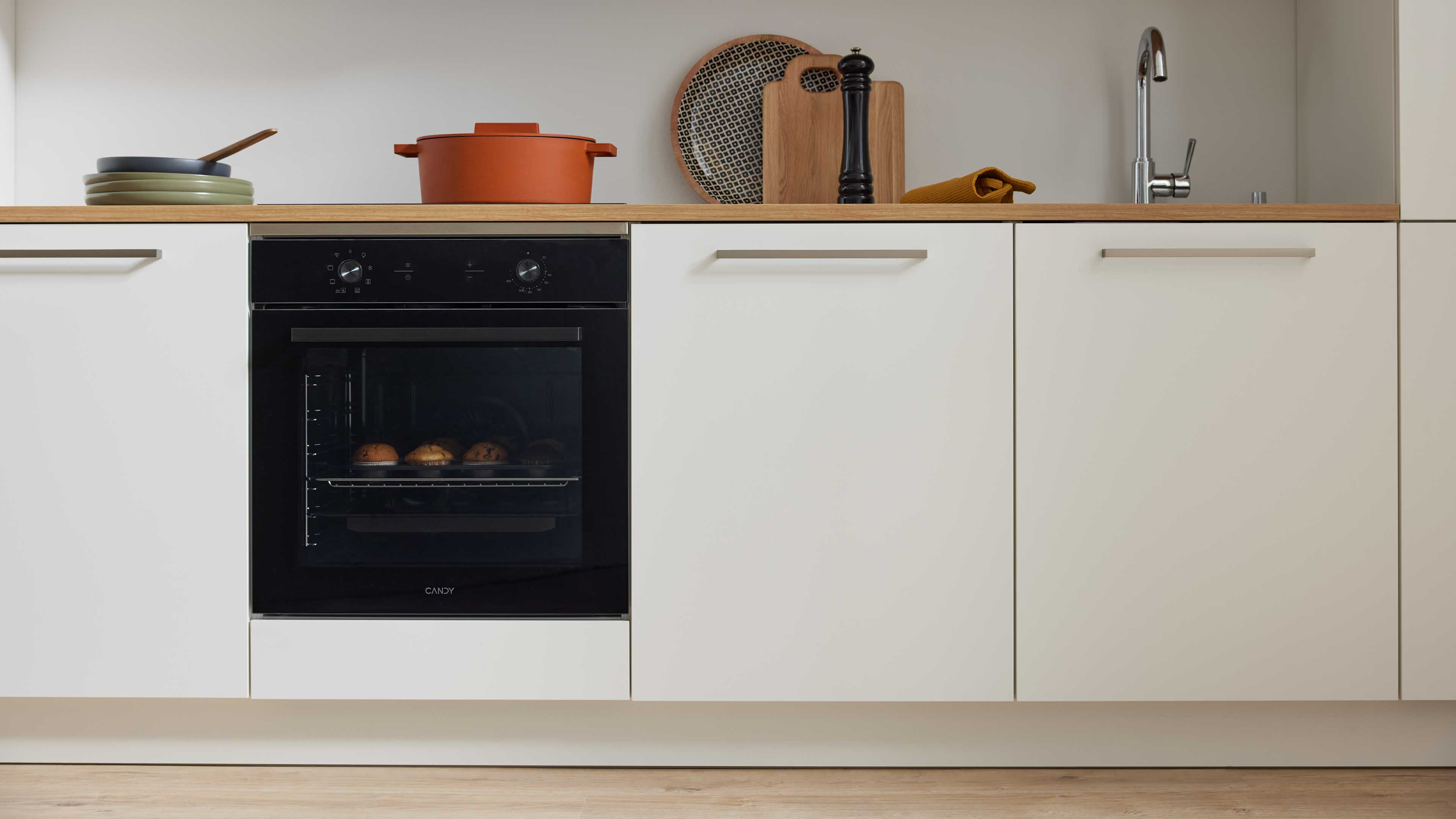Schwarzer Einbaubackofen mit sichtbaren Muffins, auf Holzarbeitsplatte Teller, Topf, Schneidebrett, Pfeffermühle und Spültisch.