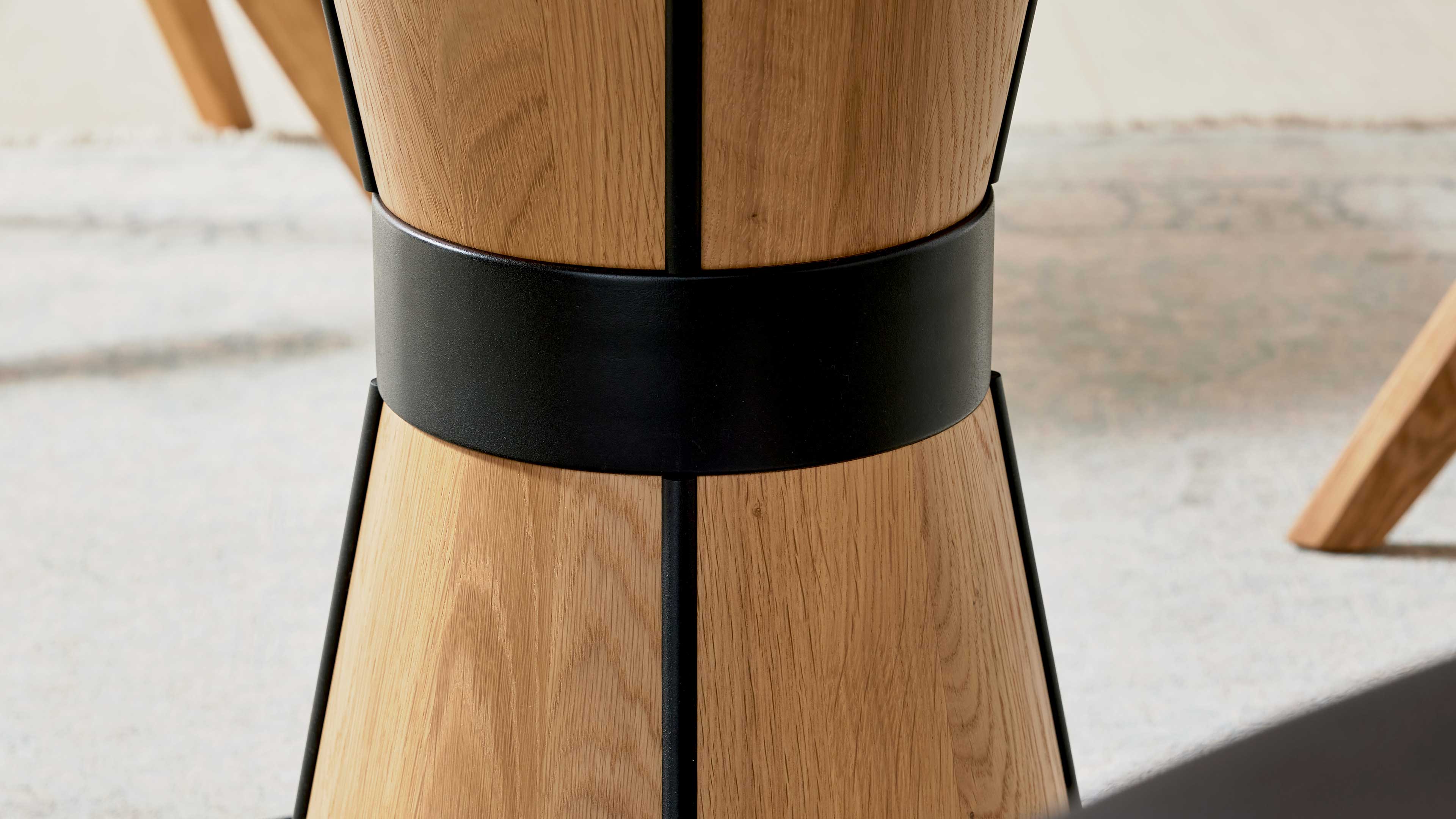 Zylindrische Tischsäule aus hellem Holz mit breitem schwarzen Metallband und dunklen senkrechten Metallstreifen, Teppichboden im Hintergrund.