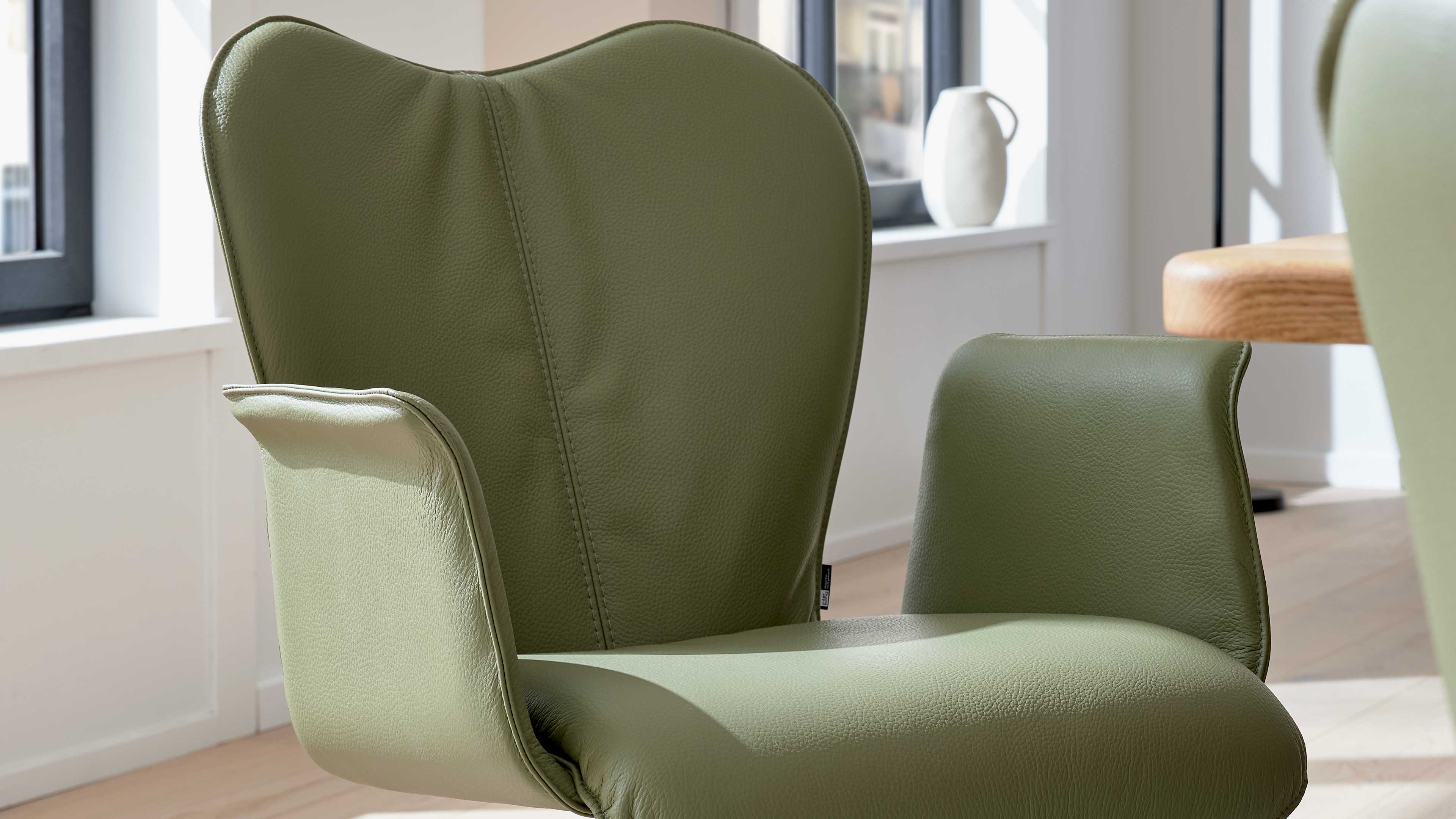 Grün bezogener Sessel mit geschwungenem Rücken, gepolsterten Armlehnen und sichtbarer Nahtstruktur, in hellem Zimmer.