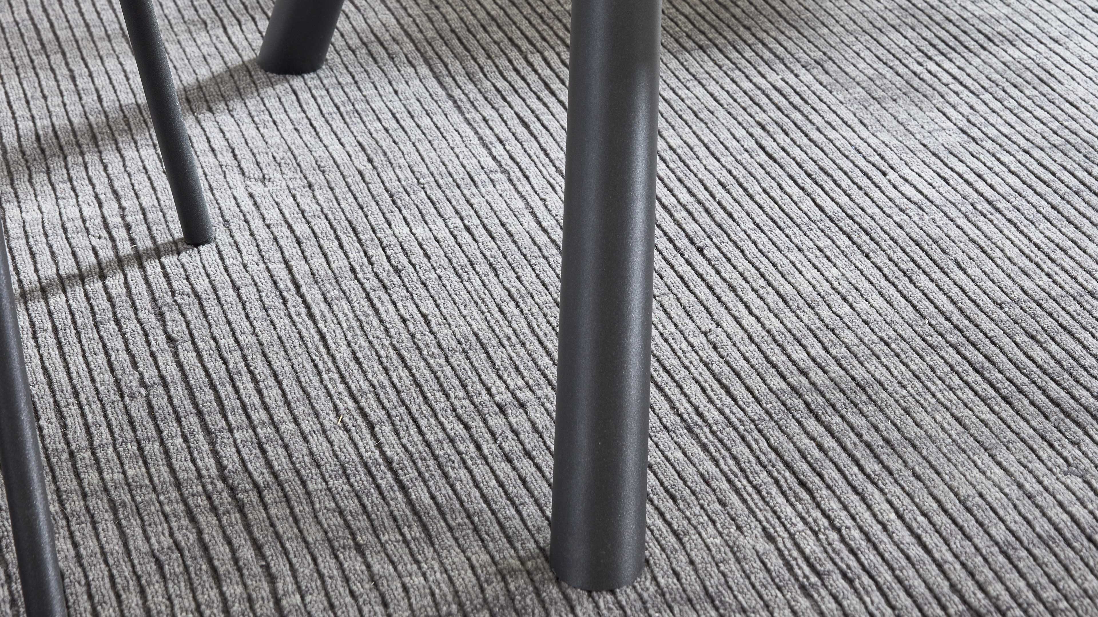 Zylindrisches dunkelgraues Metallbein im Vordergrund, zwei weitere schlanke Metallbeine links auf grauem, geripptem Teppich.