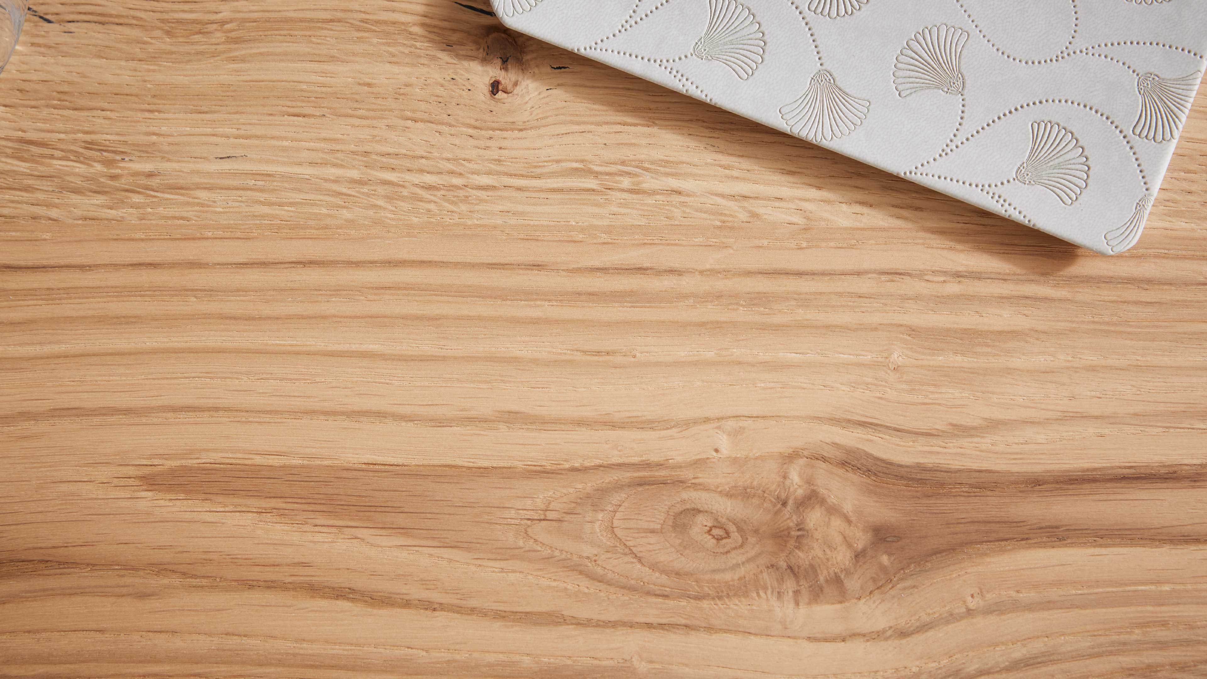 Helles Holztischblatt mit sichtbarer Maserung und Ast, oben rechts ein weißes rechteckiges Objekt mit eingeprägten Muschelmotiven.