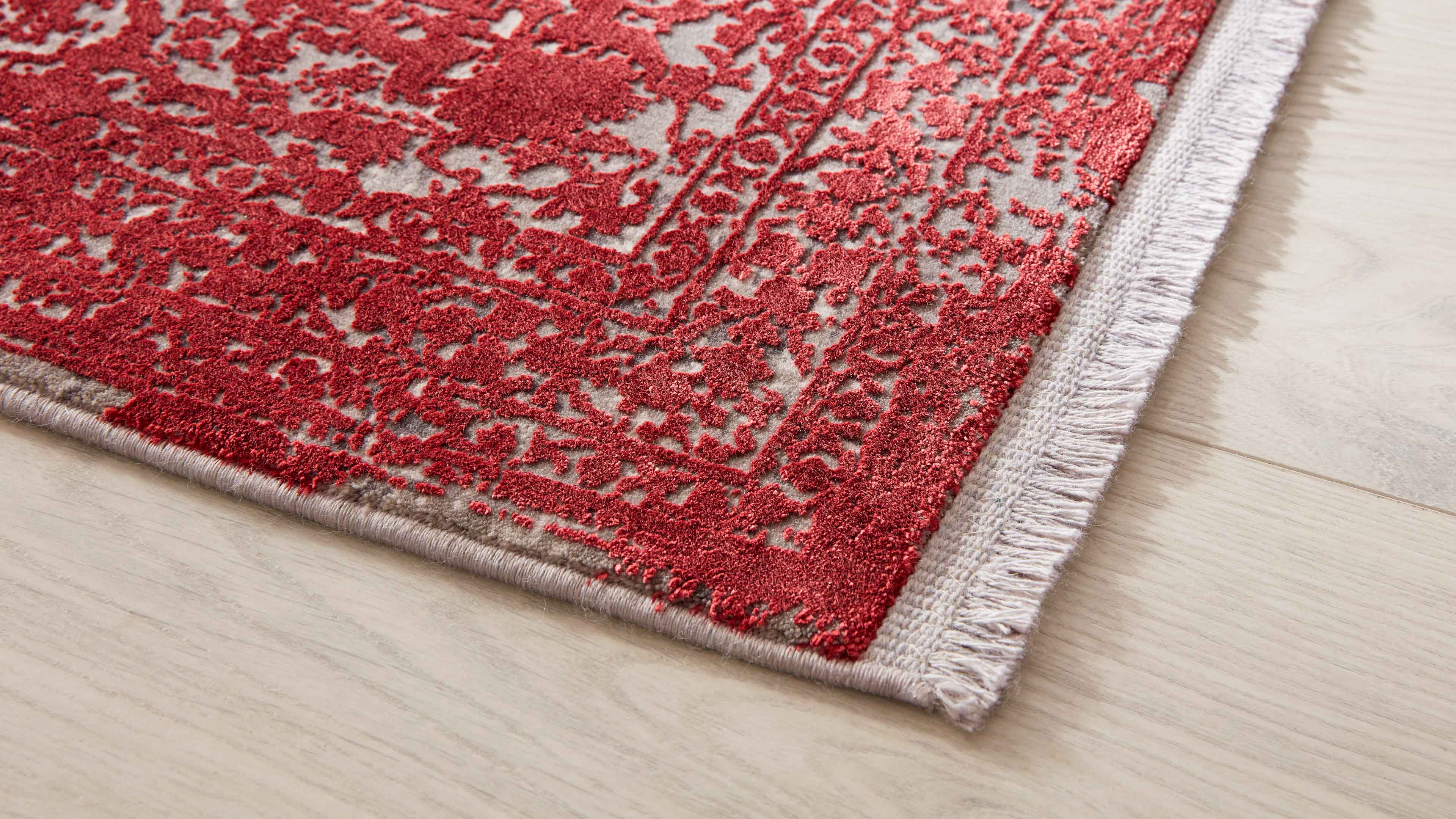Eckansicht eines Teppichs mit erhabener roter Ornamentstruktur auf hellem beigen Grund und seitlicher weißer Fransenborte auf Holzfußboden.