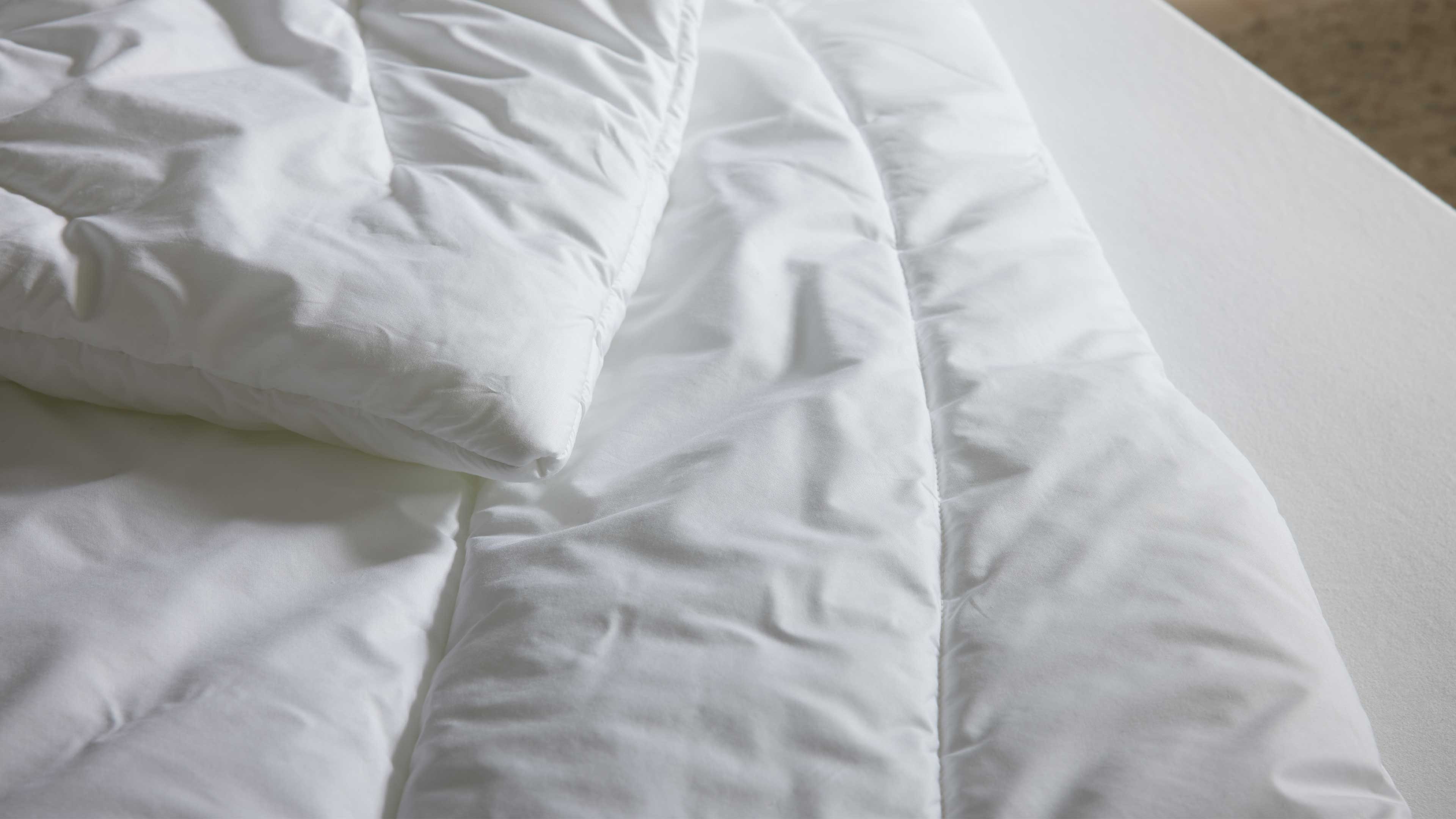 Weiße gesteppte Bettdecke mit sichtbaren Nähten und Falten auf weißem Matratzenbezug, obere Ecke umgeschlagen.